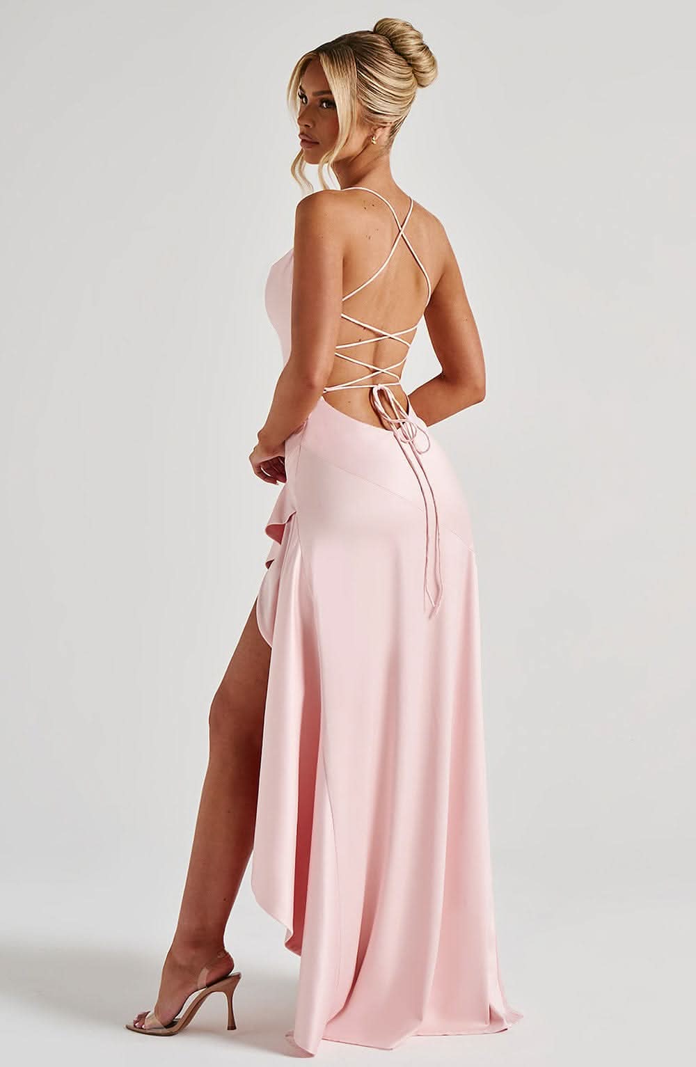 Side profile of a light pink satin maxi dress, highlighting the high-low ruffled skirt, high slit, and criss-cross back straps, worn with nude heels.