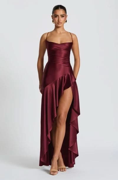 Front view of a deep burgundy satin maxi dress with spaghetti straps, a draped neckline, ruched waist, and a high-low ruffled skirt with a high slit.