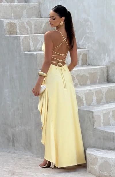 Woman in a light yellow maxi dress with criss-cross back detail and a side ruffle, posing on stone stairs.