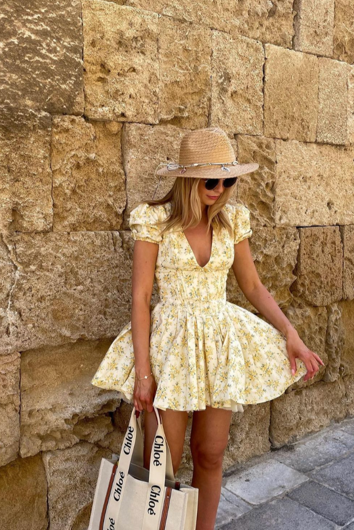 Woman in a yellow floral Delaney Pleated Mini Dress with a deep v-neckline and pleated skirt detail.