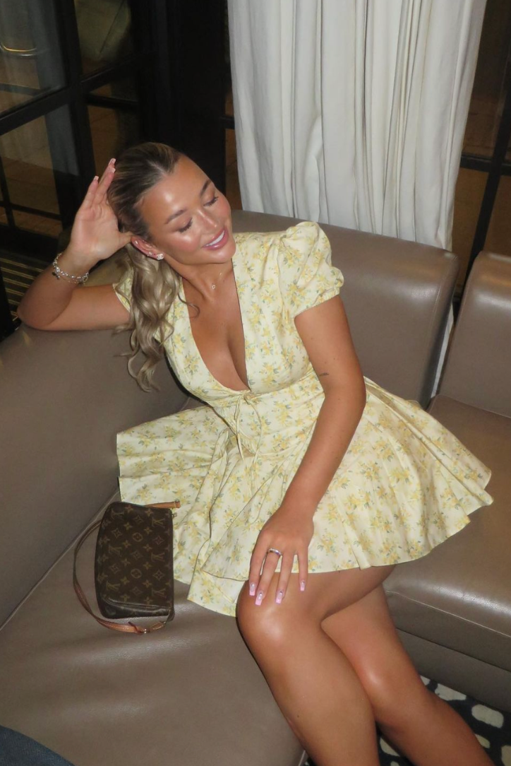 Yellow floral Delaney mini dress with a plunging neckline and cinched waist, sitting casually on a brown sofa.