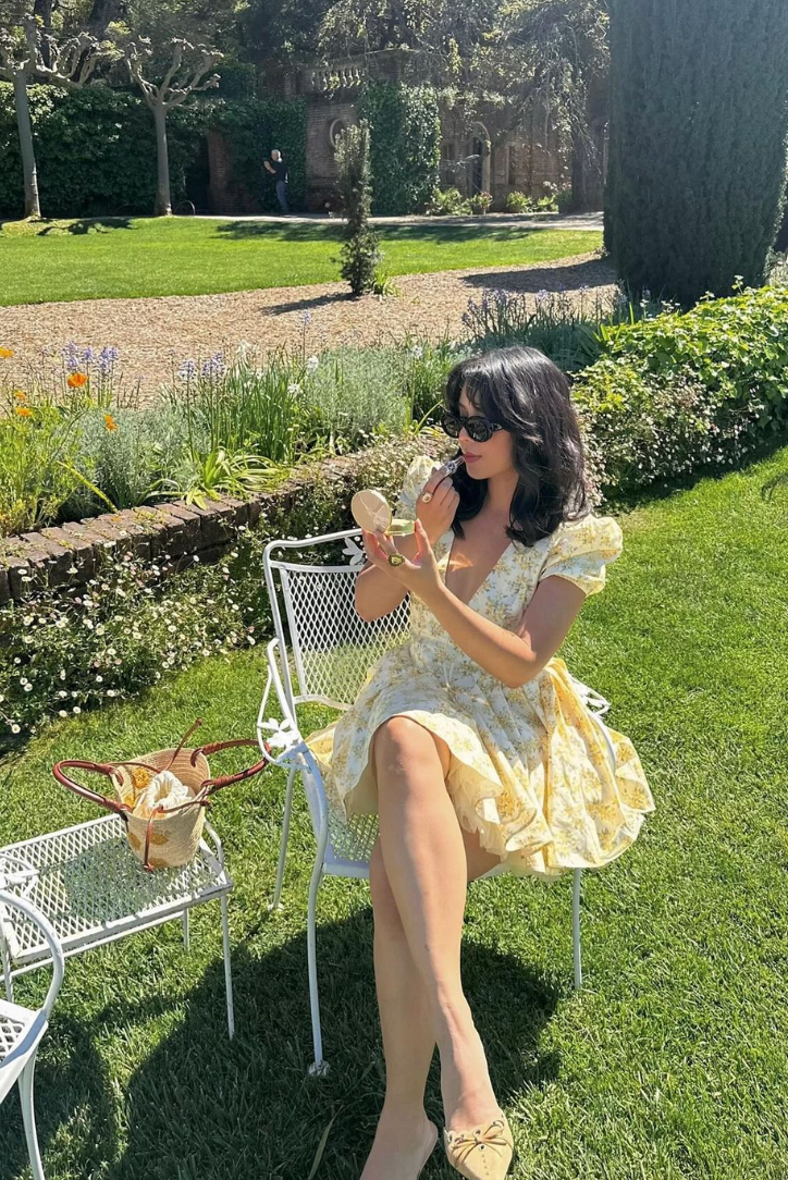 Yellow floral Delaney mini dress with puff sleeves, worn with black sunglasses and cream bow slides, sitting in a garden.