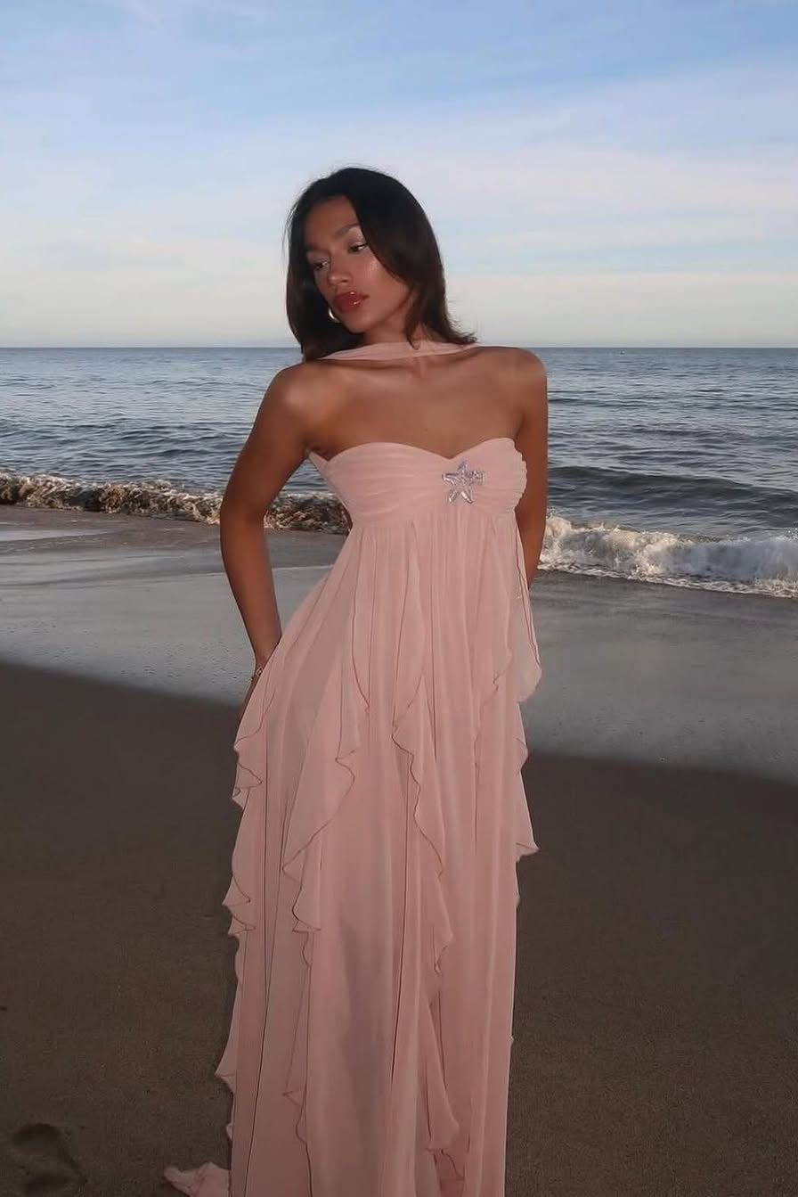 Pale pink ruched maxi dress with a strapless sweetheart neckline and delicate ruffles, worn on a beach at sunset.