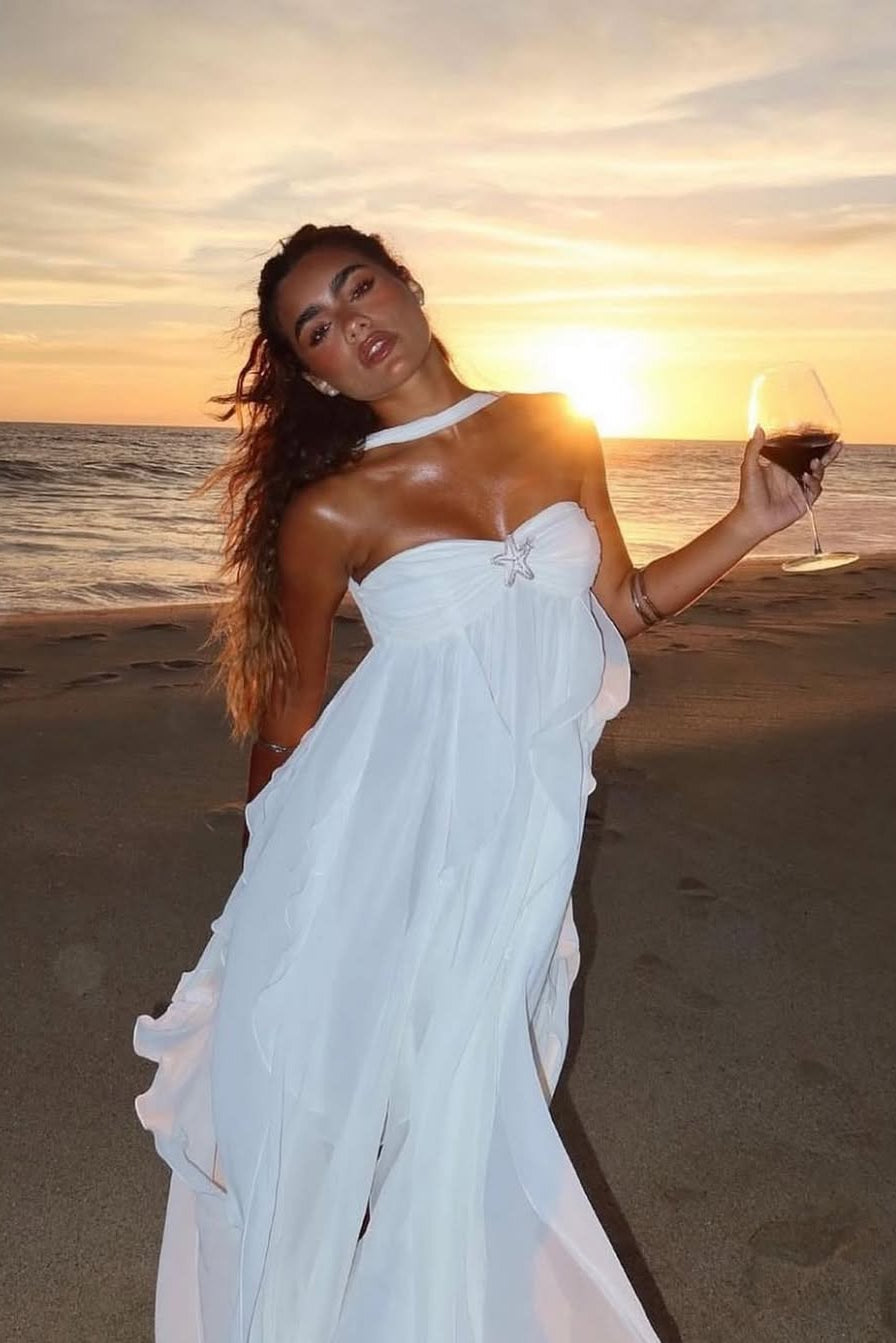 White ruched maxi dress with a strapless neckline, halter tie, and layered ruffled skirt, on a beach at sunset.