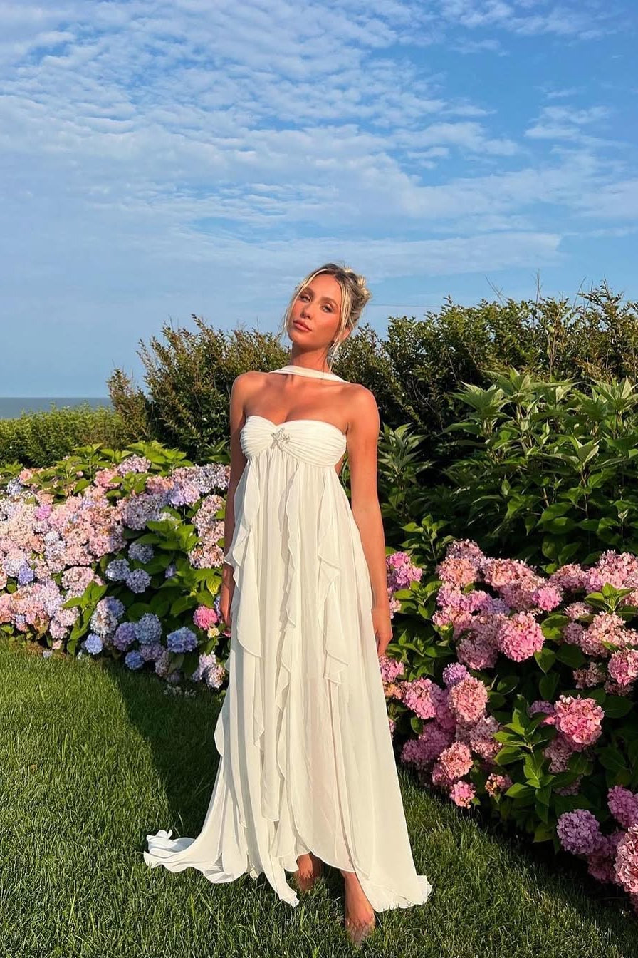 White ruched maxi dress with a strapless top and flowing ruffled skirt, standing in a garden with pink flowers.
