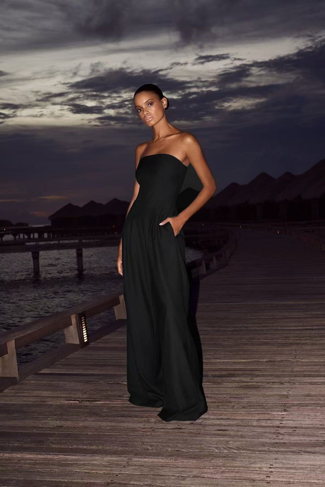 Full-length view of a woman wearing a navy blue strapless wide-leg jumpsuit with side pockets and a white crossbody bag.