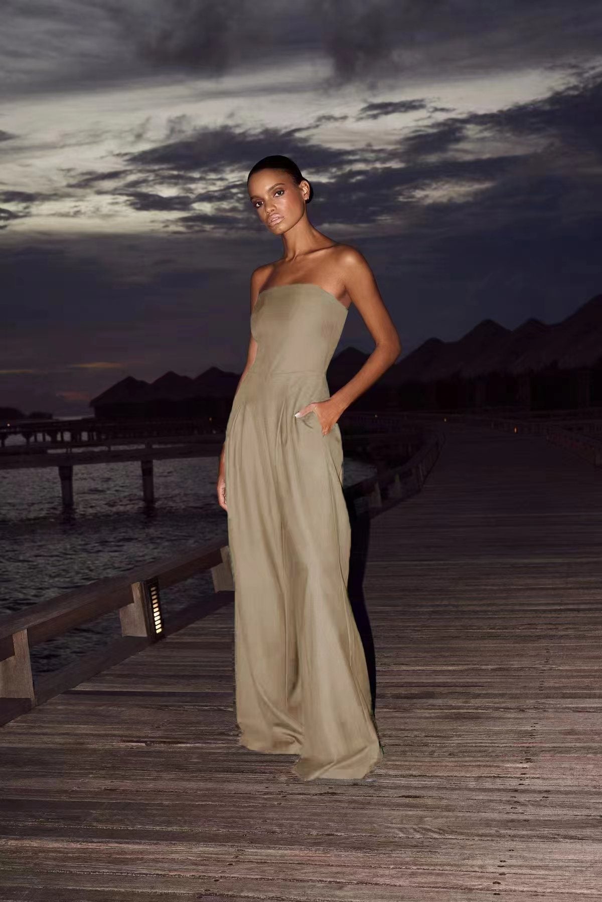 Woman on a wooden pier at dusk, wearing a light olive green strapless wide-leg jumpsuit with side pockets.