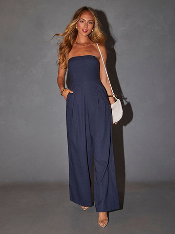 Full-length view of a woman wearing a navy blue strapless wide-leg jumpsuit with side pockets and a white crossbody bag.