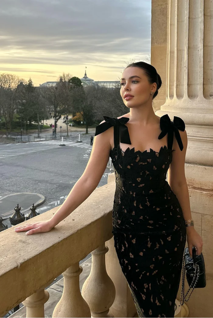 Black lace Elle Bow Maxi Dress with structured shoulder bows, worn by a woman on a balcony overlooking a city.