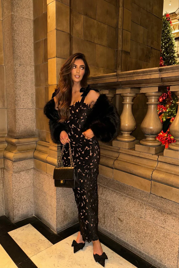 Woman wearing a black lace Elle Bow Maxi Dress with a black faux fur jacket and bow-detail heels.