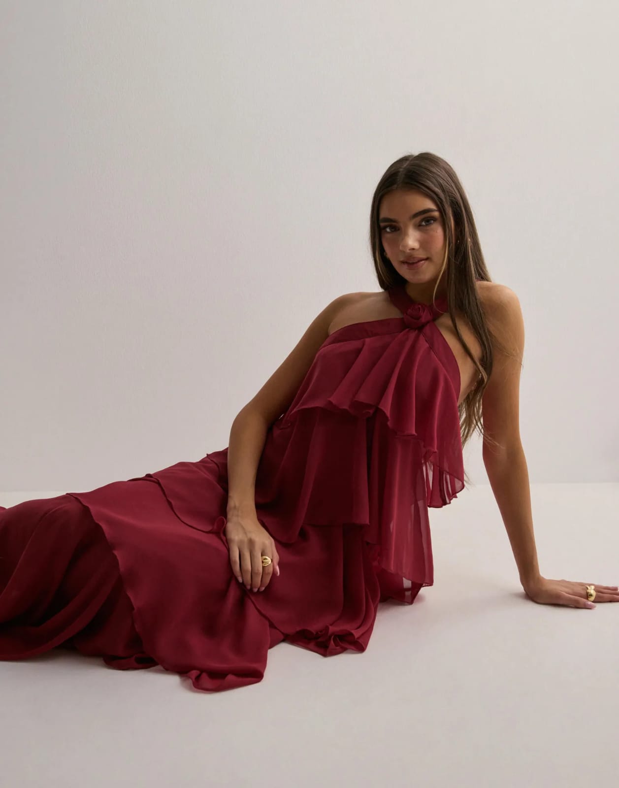 Woman reclining in a deep red ruffle maxi dress, featuring a halter neckline and tiered skirt, with gold rings.