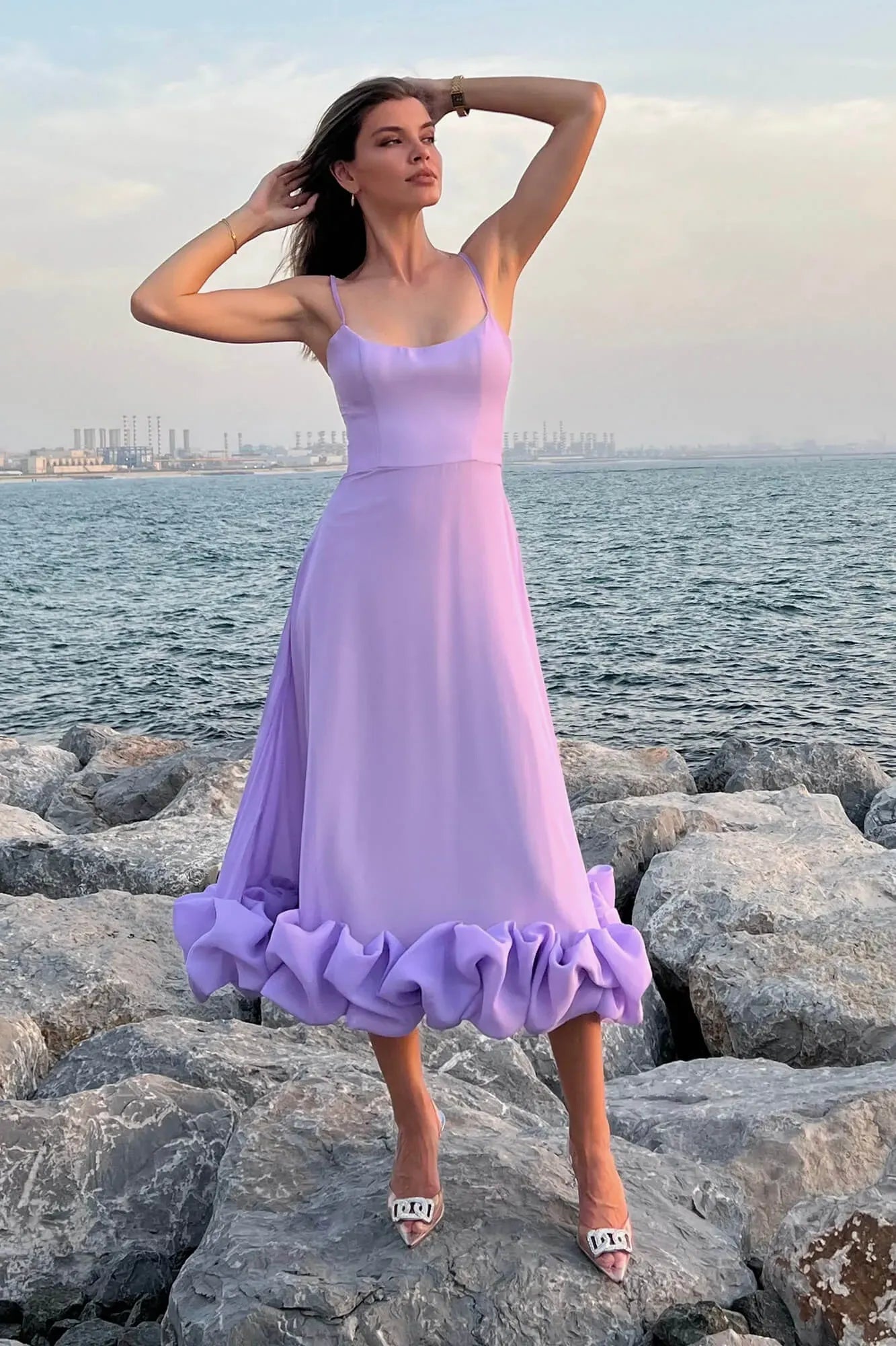 Lavender midi dress with thin straps and a large ruffled hem, worn with high heels by the ocean.