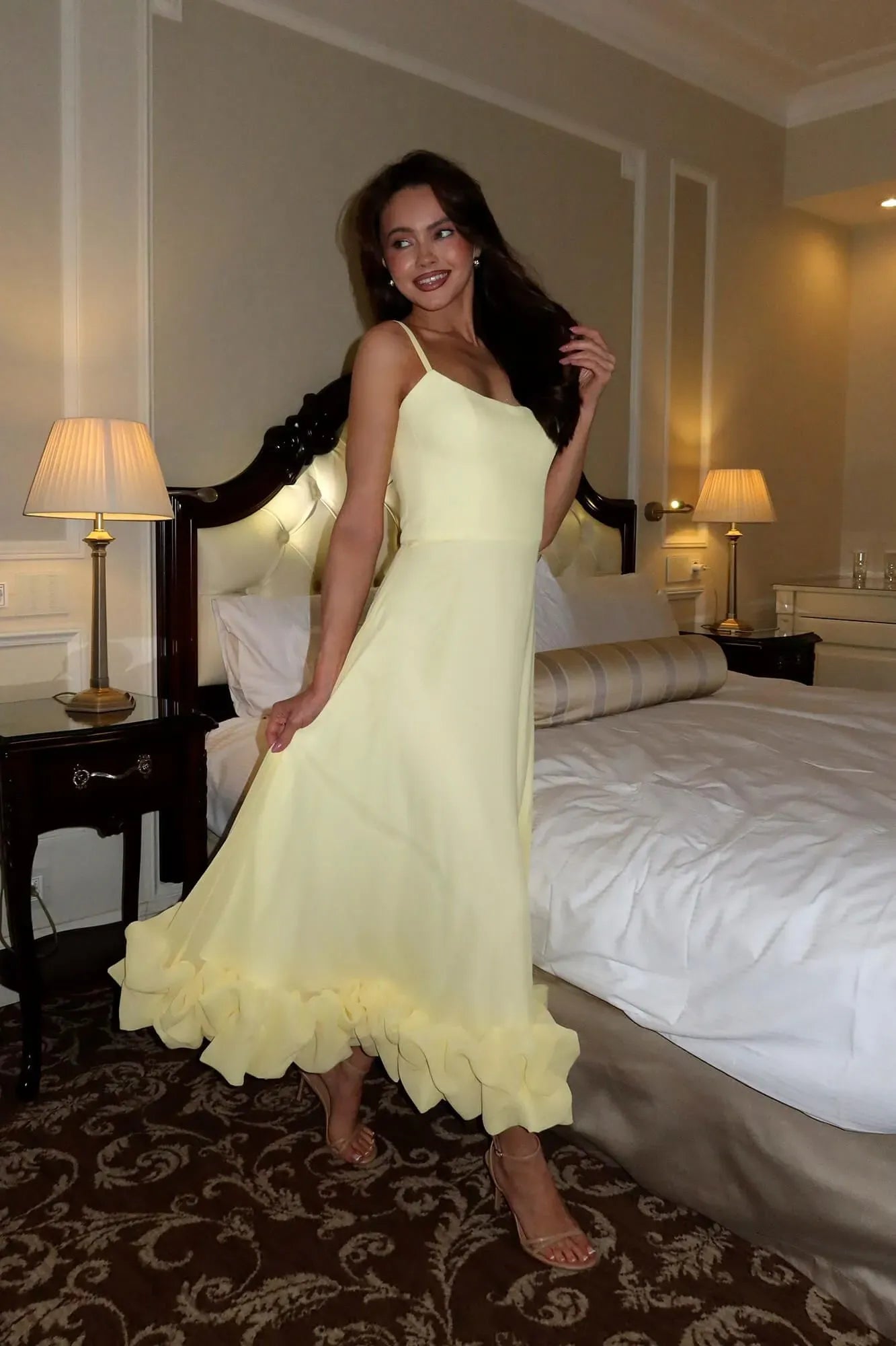 Light yellow Emmeline ruffle midi dress with spaghetti straps and a tiered ruffle hem, in a hotel room setting.