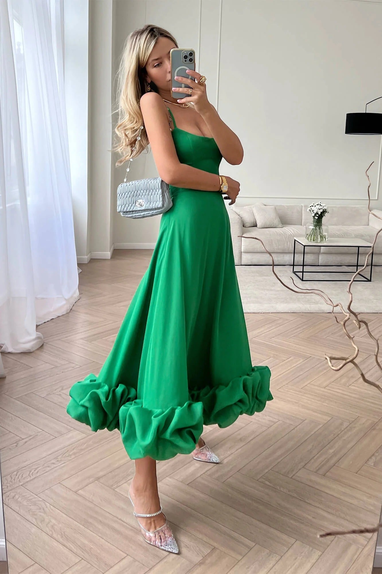 Full-length emerald green Emmeline Ruffle Midi Dress with spaghetti straps and a voluminous ruffled hem, worn with clear pointed heels and a light blue quilted purse.