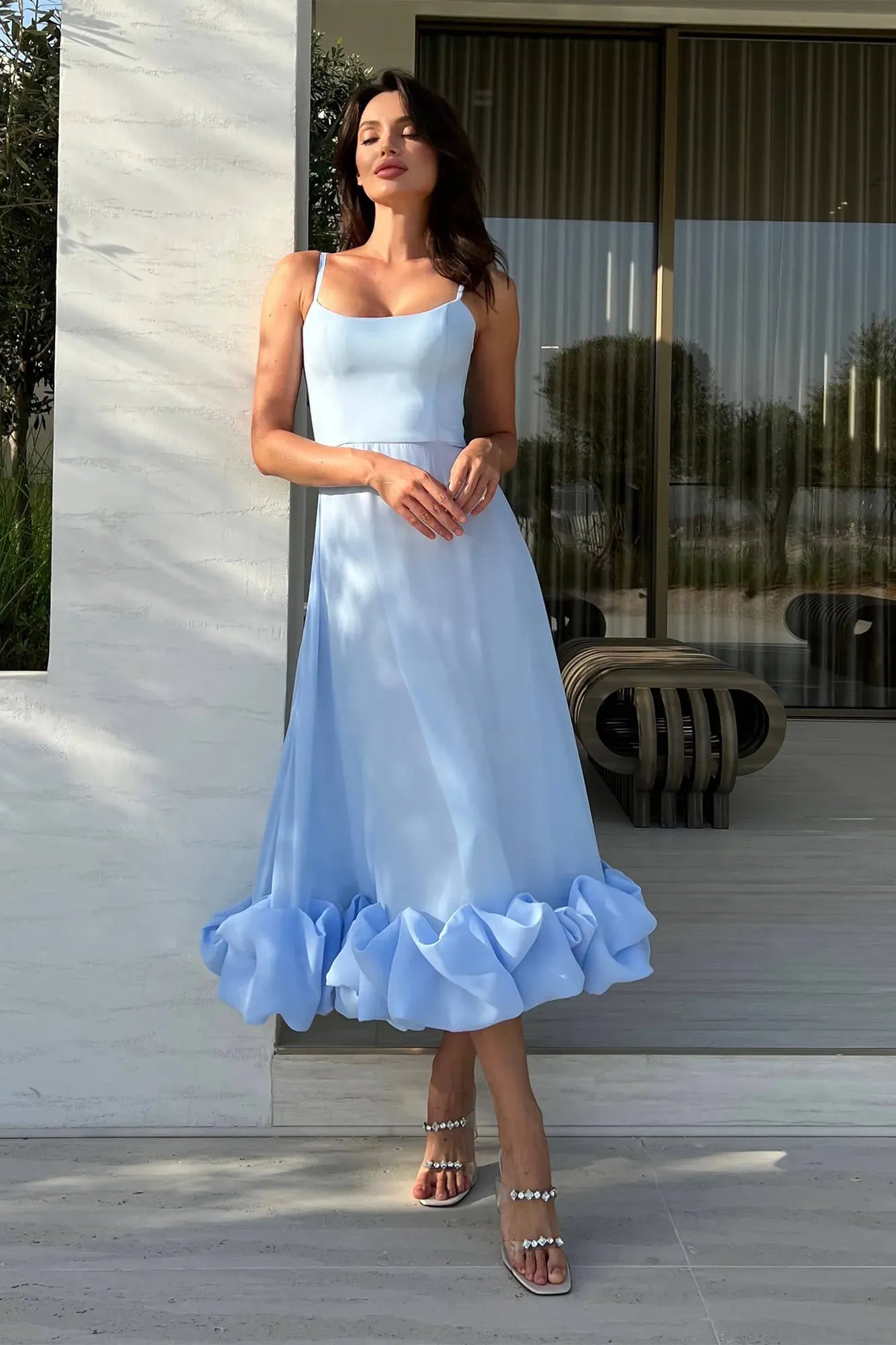 Front view of a light blue midi dress with spaghetti straps and voluminous ruffled hem, with clear heels.