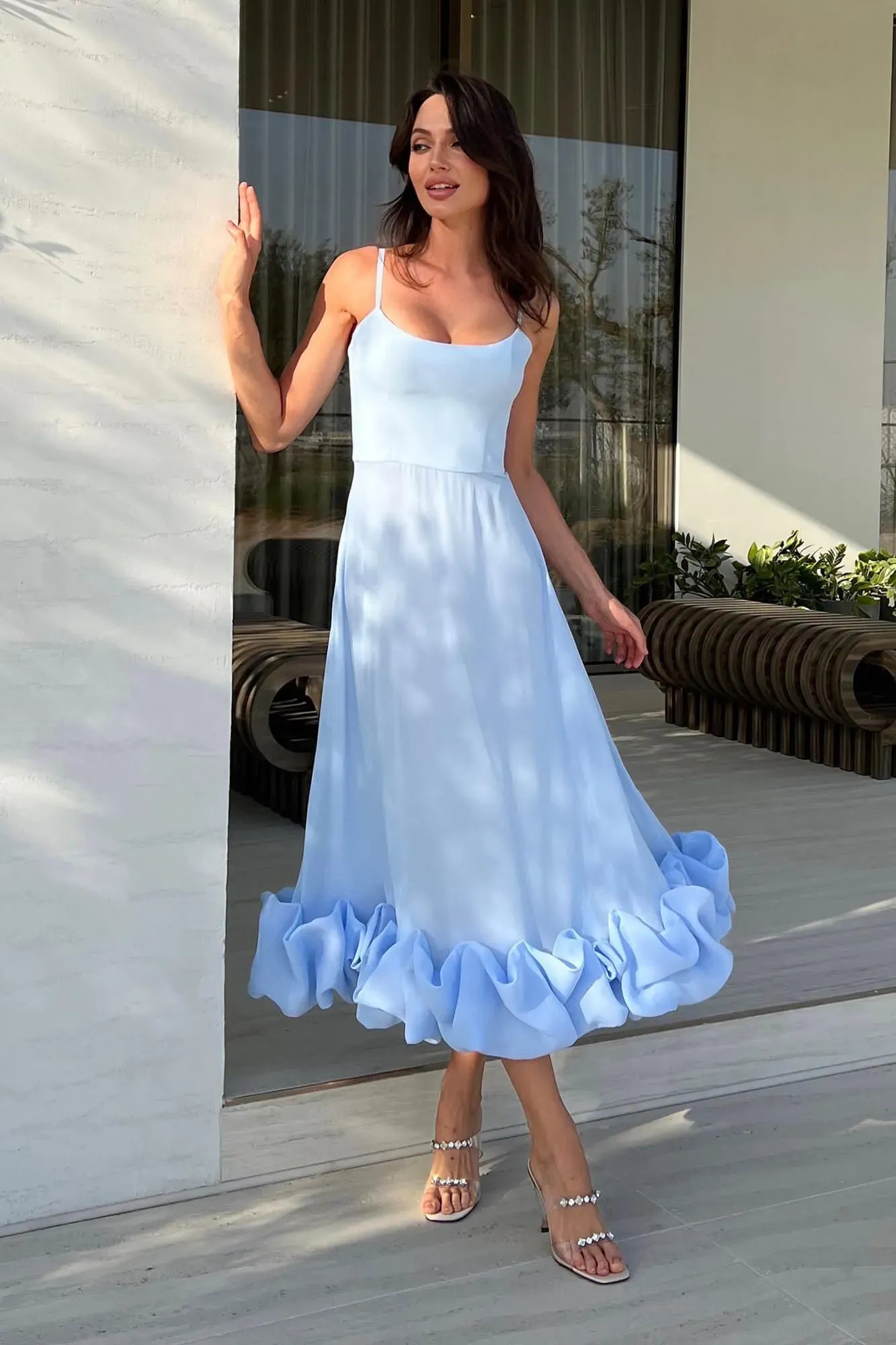 Full body shot of a light blue midi dress with thin straps and a large ruffled hem, worn outdoors.