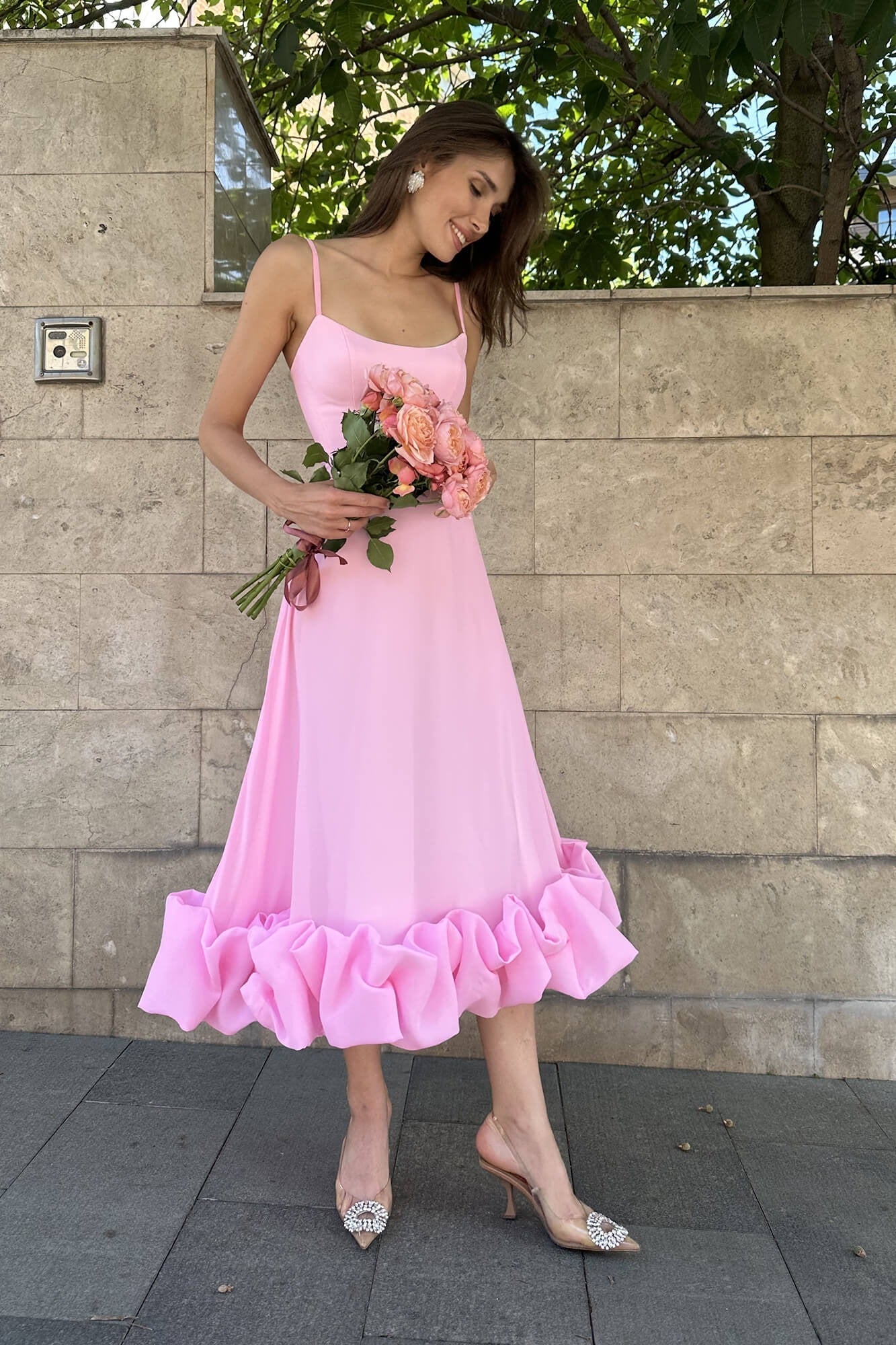 Pink midi dress with spaghetti straps and a ruffled hem, holding a bouquet of pink roses.