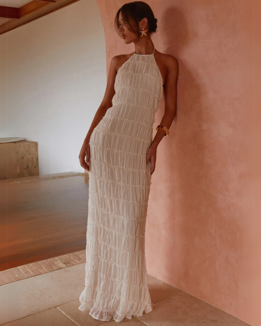 Everly textured maxi dress in off-white, featuring a halter neck and ruched detailing, styled on a model leaning against a pink wall.
