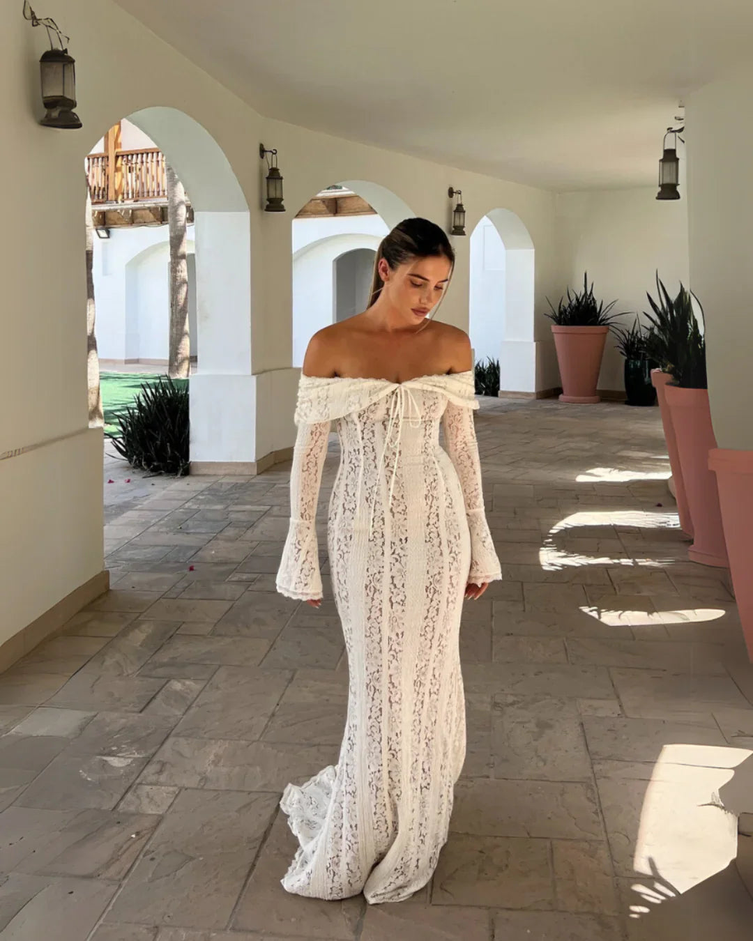 Off-the-shoulder white lace maxi dress with long bell sleeves and a tie-front detail, walking on a patio.