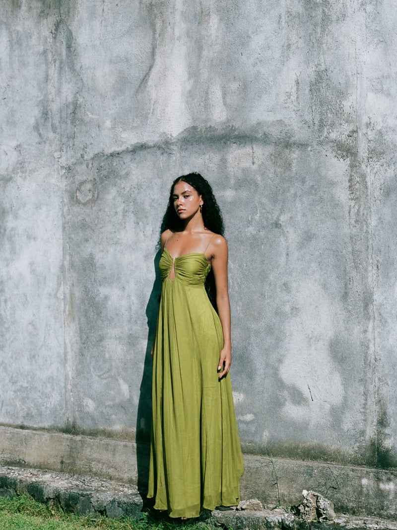 Woman in a long olive green maxi dress with keyhole detail, standing against a textured grey wall in natural light.