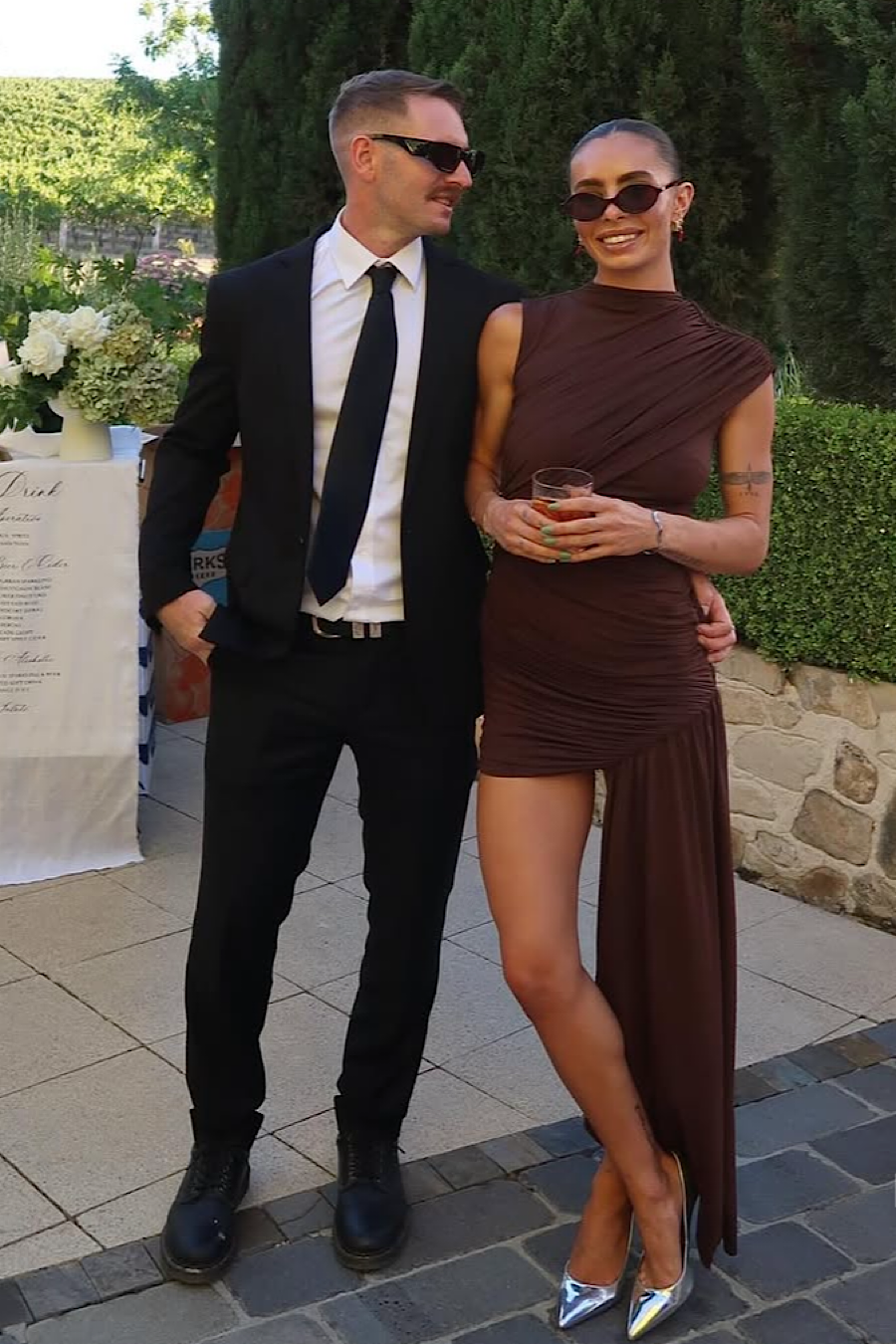 Outdoor shot of a woman in a brown ruched mini dress and silver heels, next to a man in a black suit.