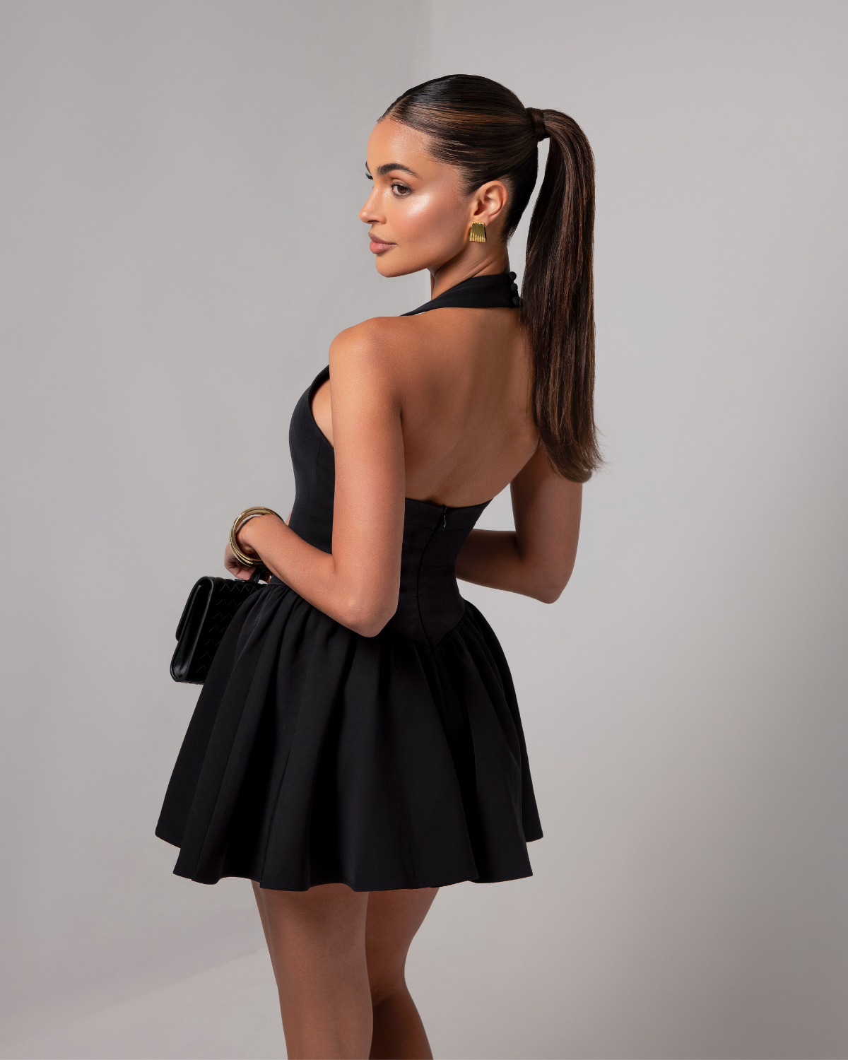 Rear view of the black Hailey halter mini dress, highlighting the halter neckline and a-line skirt, holding a black clutch.