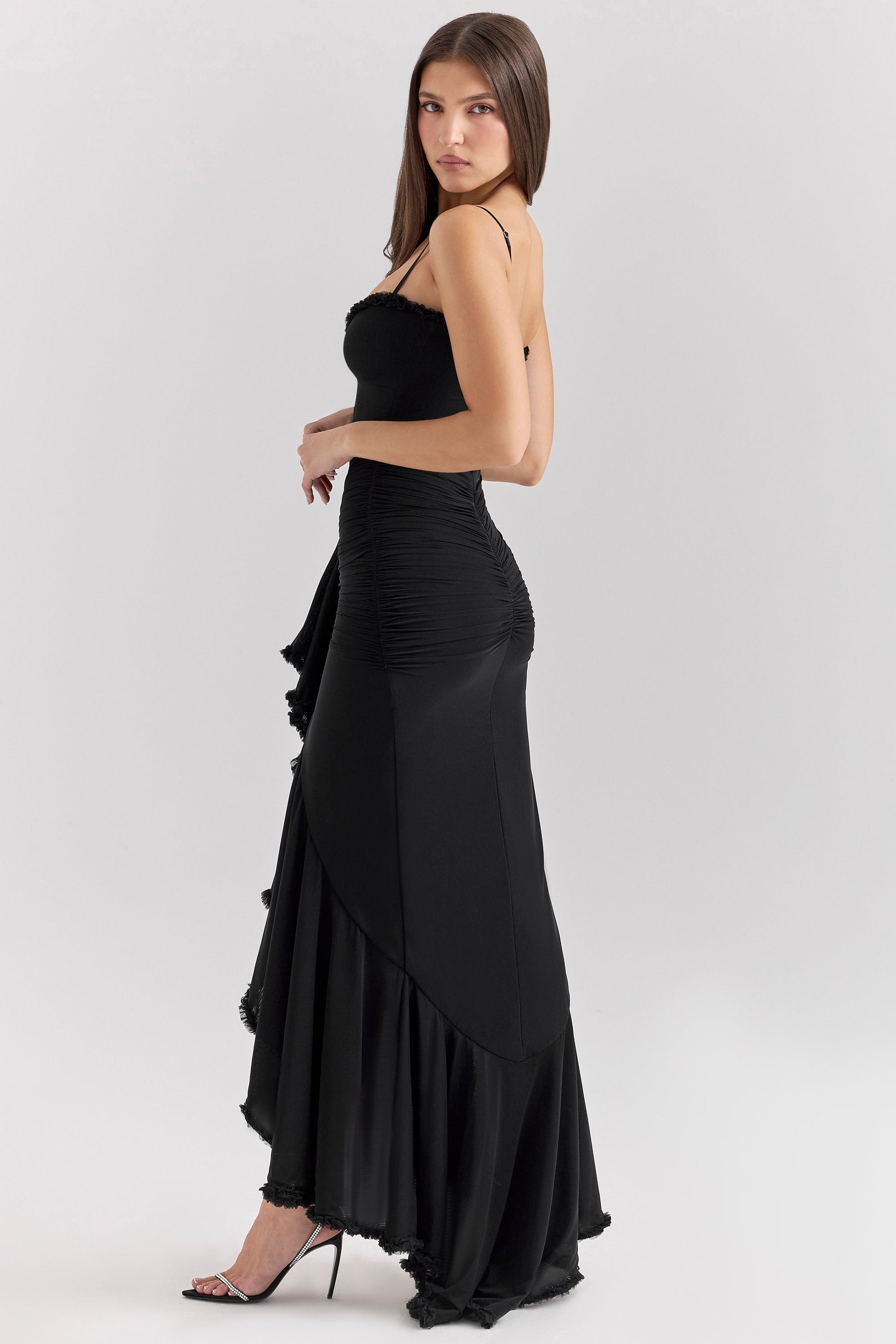 Side profile of a woman wearing a black Isla Ruffle Maxi Dress, highlighting the cascading ruffle and fitted silhouette with heels.