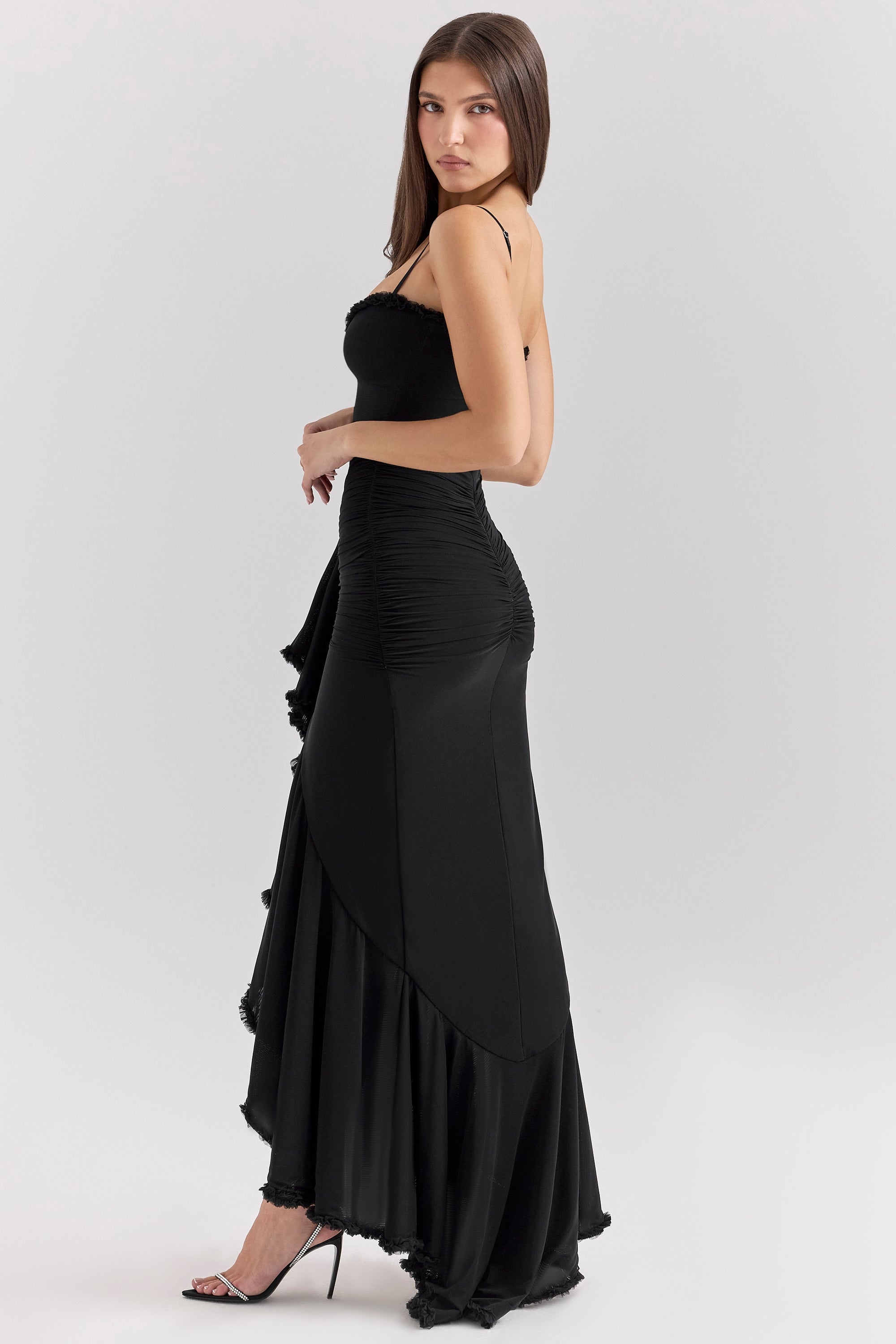 Side profile of a woman wearing a black Isla Ruffle Maxi Dress, highlighting the cascading ruffle and fitted silhouette with heels.