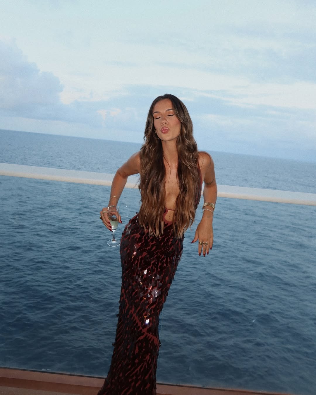 Full length view of the burgundy sequin maxi dress with a deep v-neck and side cut-outs, worn on a balcony at sunset.