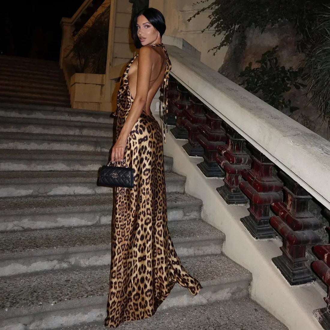 Full-length Jillian leopard print maxi dress with halter neckline and deep V-neck, cinched waist, flowing skirt, worn on stairs.