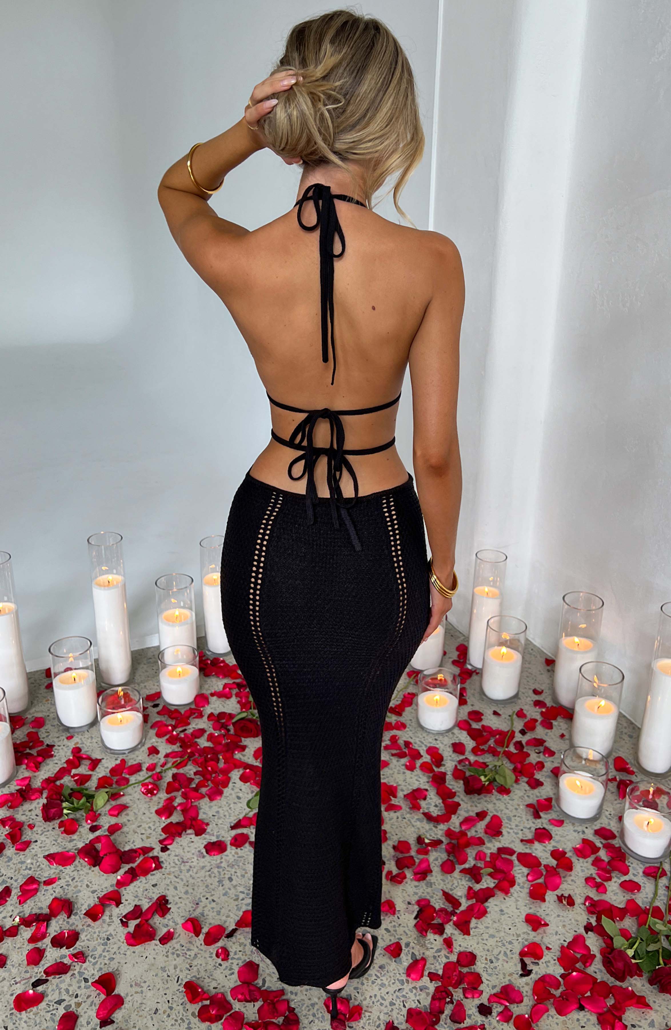 Back view of the black Kara Cutout Maxi Dress, revealing the tie-back halter and open back design, with candles and rose petals on the floor.