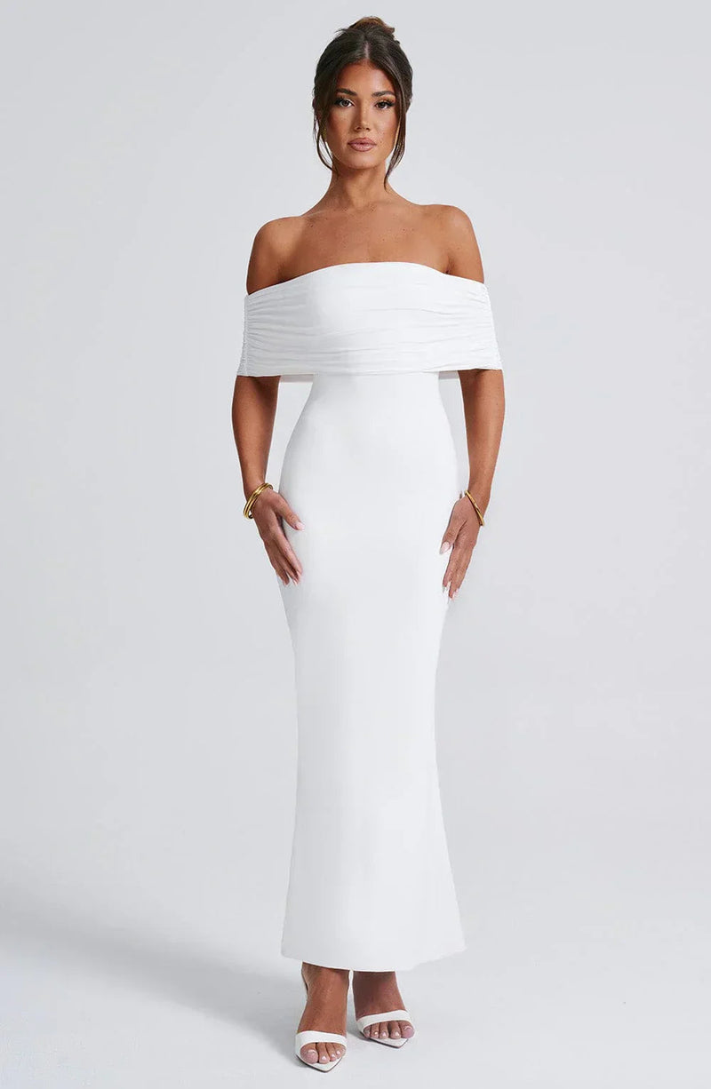 Front view of a women's elegant white Karina maxi dress, featuring an off-the-shoulder pleated neckline and a fitted silhouette, with gold bracelets.