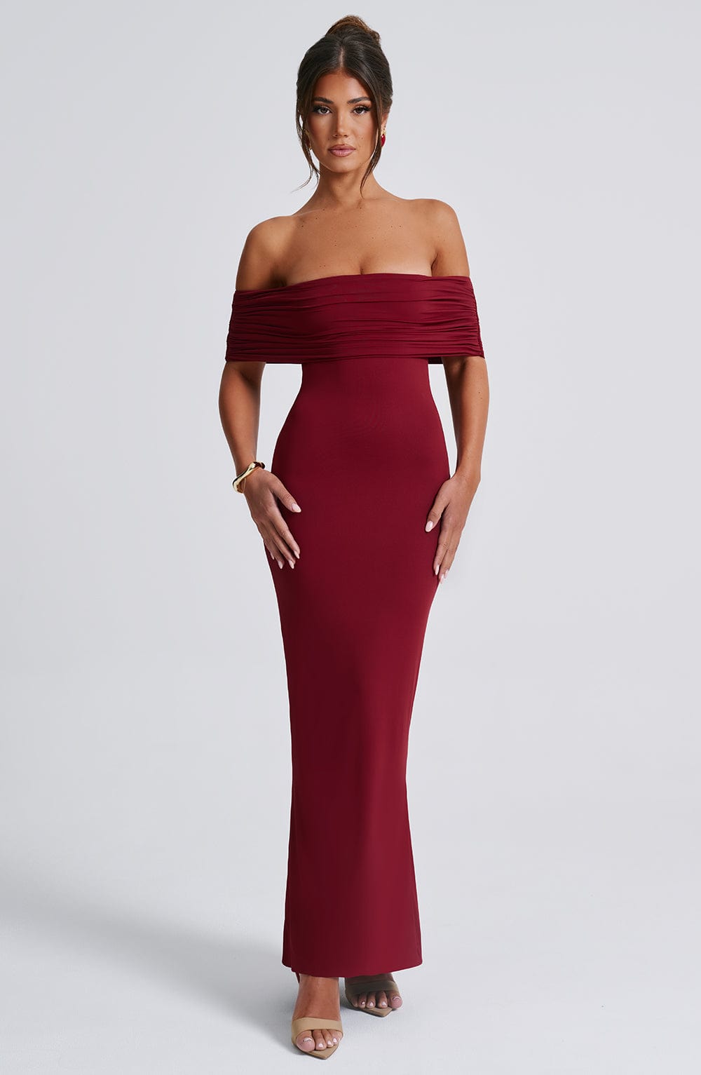 Front view of a women's deep red Karina maxi dress, showcasing an off-the-shoulder pleated bodice and a long, flowing skirt, with gold bracelet.