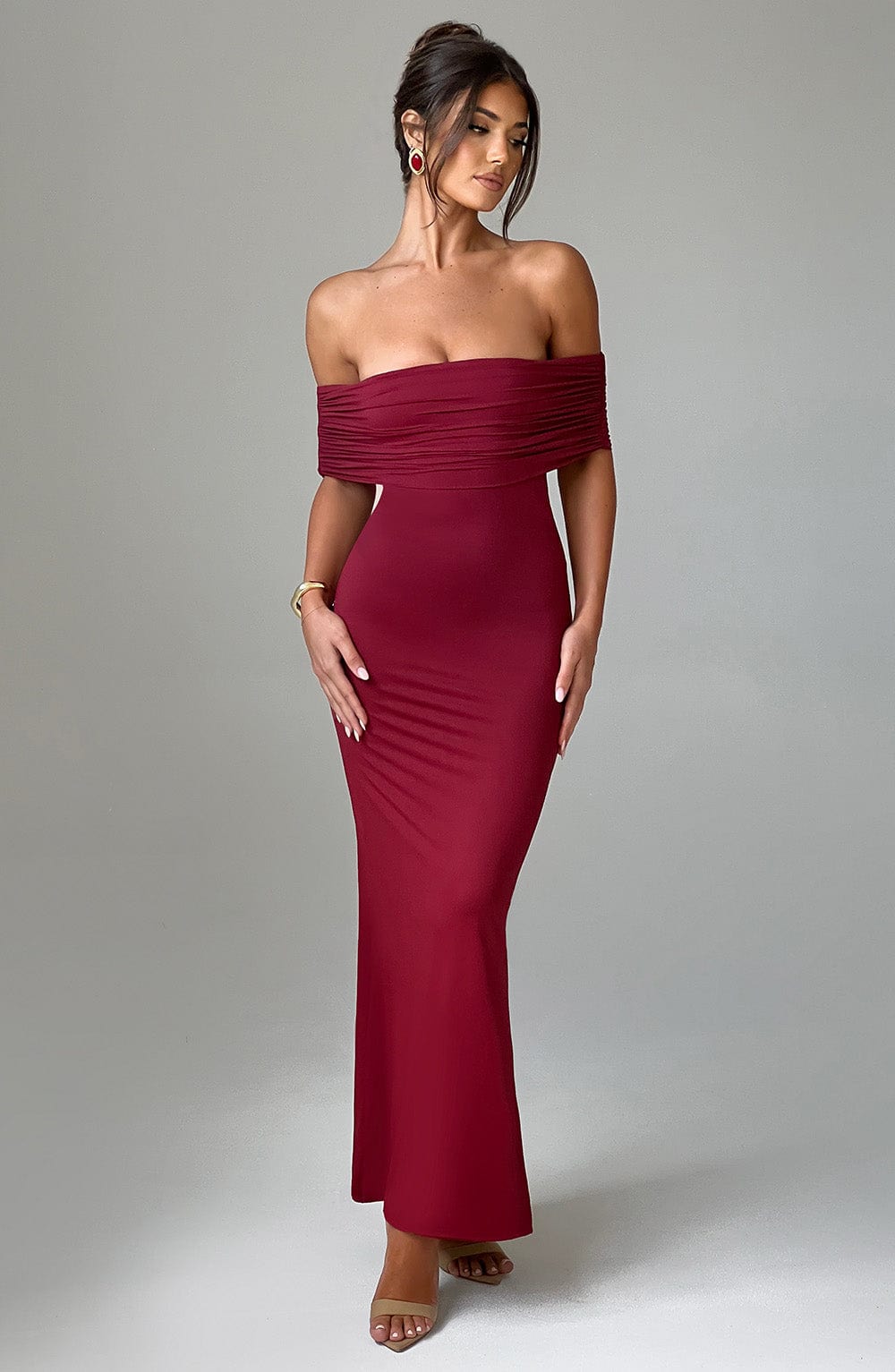 Angled front view of a women's burgundy Karina maxi dress, featuring off-the-shoulder pleats and a fitted silhouette, paired with gold earrings and bracelet.