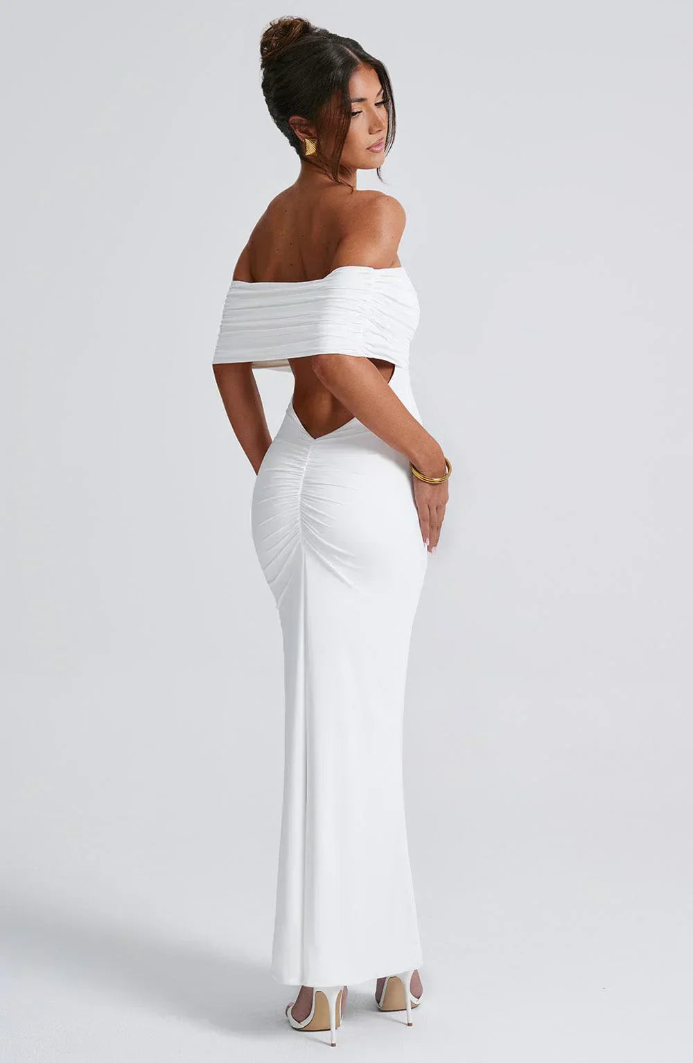 Back view of a women's white Karina backless maxi dress with off-the-shoulder draping and a ruched accent on the lower back, with gold bracelet.