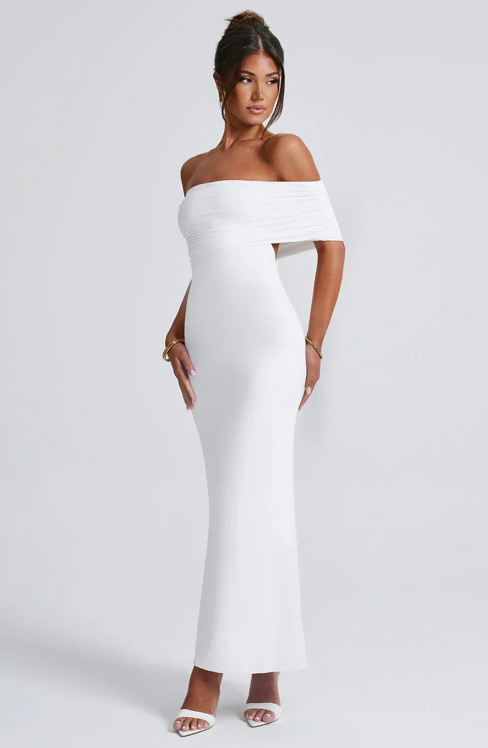 Side view of a women's white Karina maxi dress, highlighting the off-the-shoulder pleated design and elegant fitted shape, with gold bracelets.