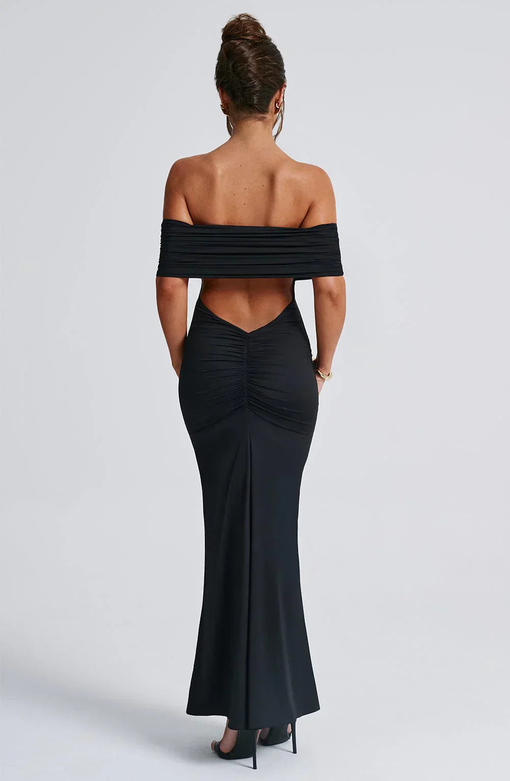 Back view of a women's black Karina backless maxi dress, featuring off-the-shoulder pleats and a gathered detail on the lower back, with gold bracelet.