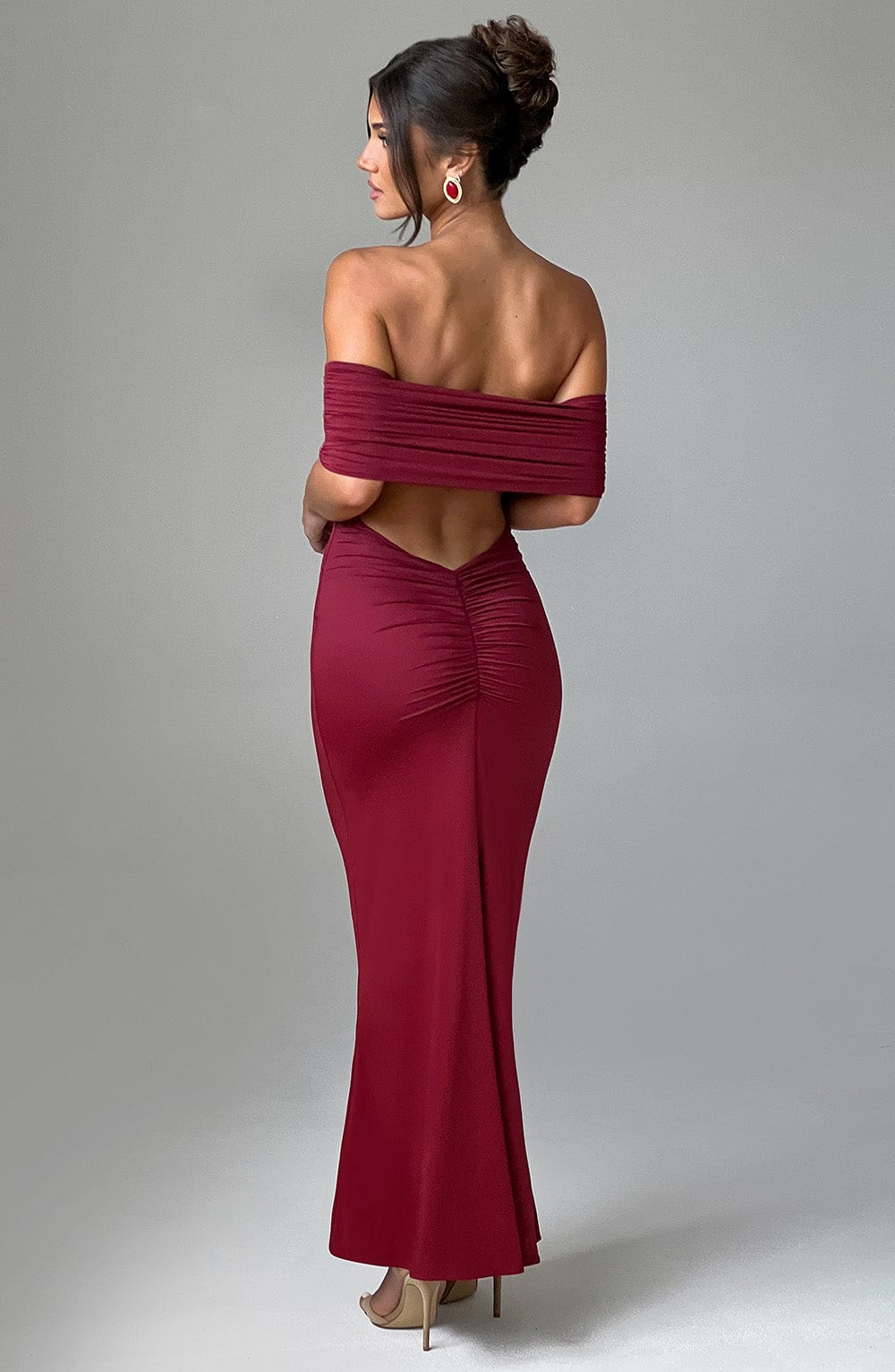 Back view of a women's rich burgundy Karina backless maxi dress with off-the-shoulder pleated detailing and ruched lower back, paired with gold heels.