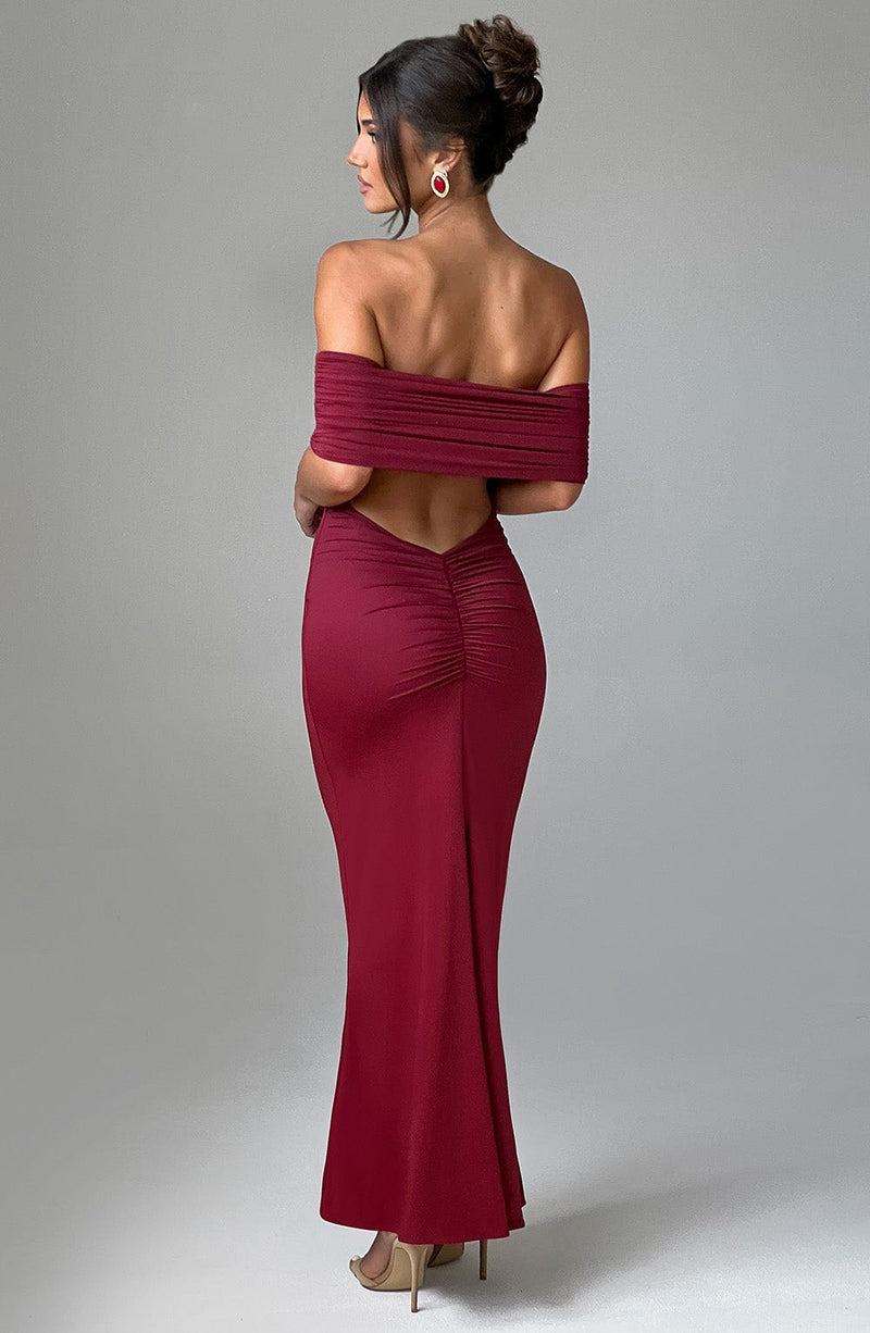 Back view of a women's rich burgundy Karina backless maxi dress with off-the-shoulder pleated detailing and ruched lower back, paired with gold heels.