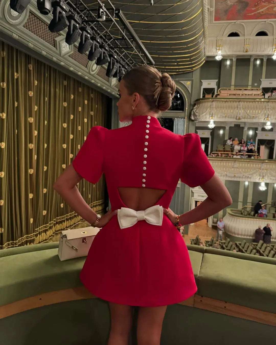 Back view of red mini dress with white bow and button details, standing on a balcony overlooking an auditorium.