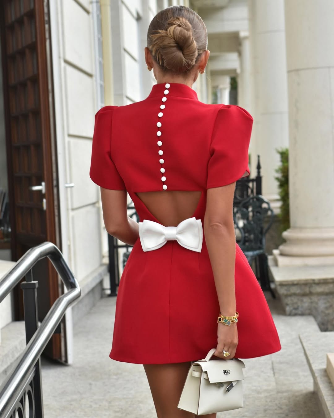 Back view of red mini dress with white bow, high collar, and white buttons down the back, holding a white handbag.