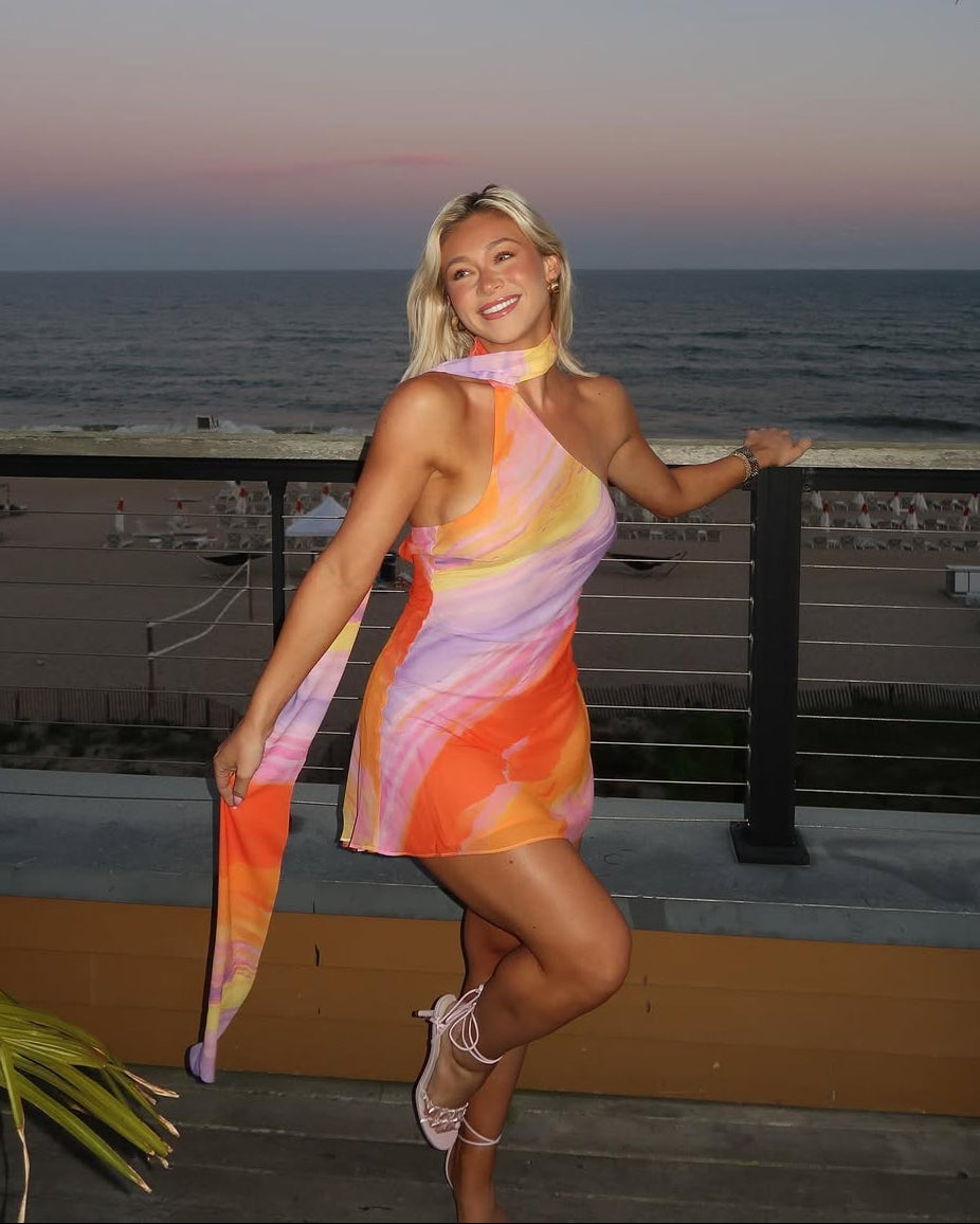 Front view of a vibrant orange, pink, and yellow abstract mini dress with a one-shoulder design and flowing scarf detail, worn with strappy heels.