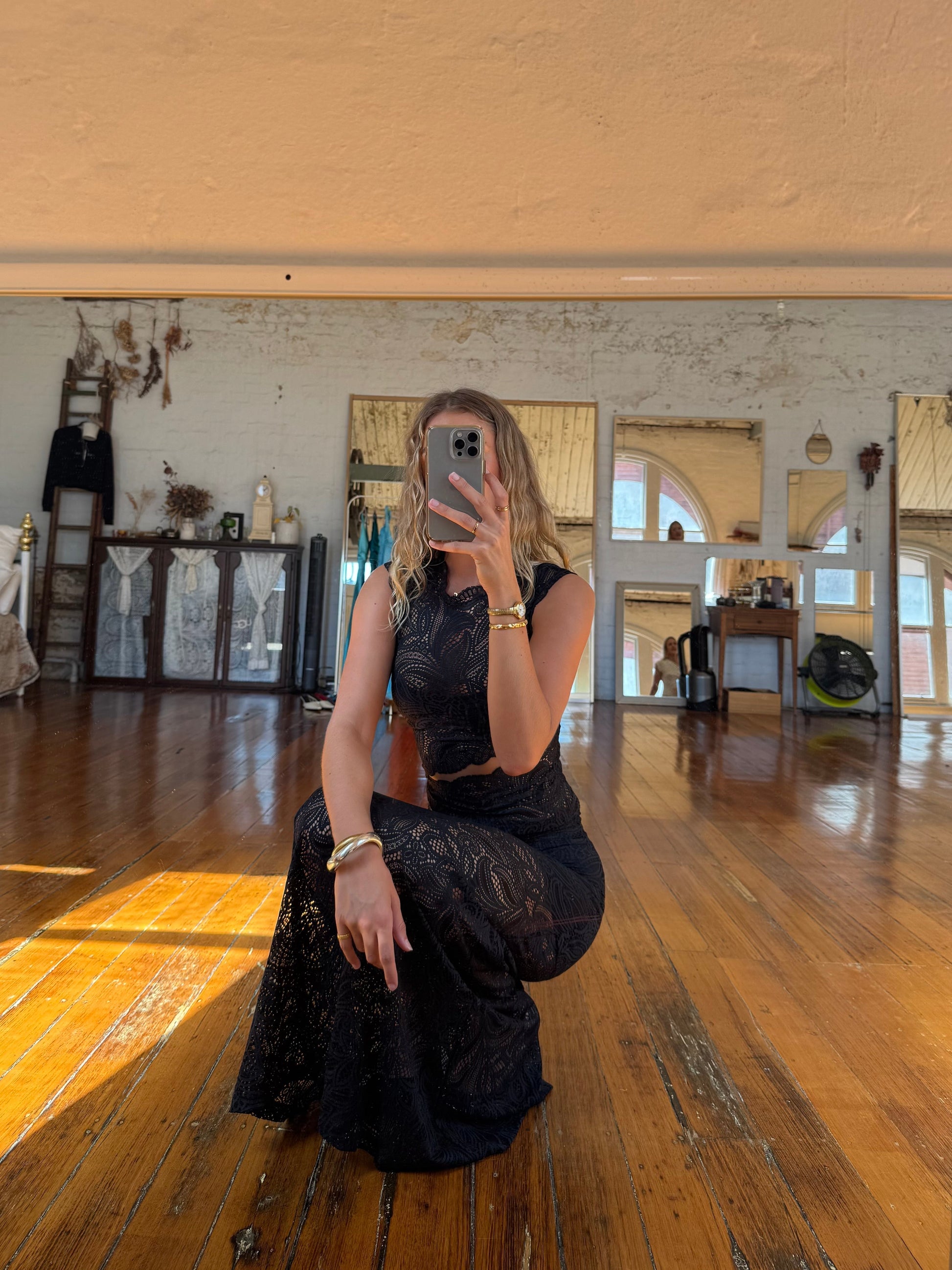 Woman in a black lace two-piece set, featuring a sleeveless crop top and a maxi skirt, posing in a well-lit room with wooden floors.