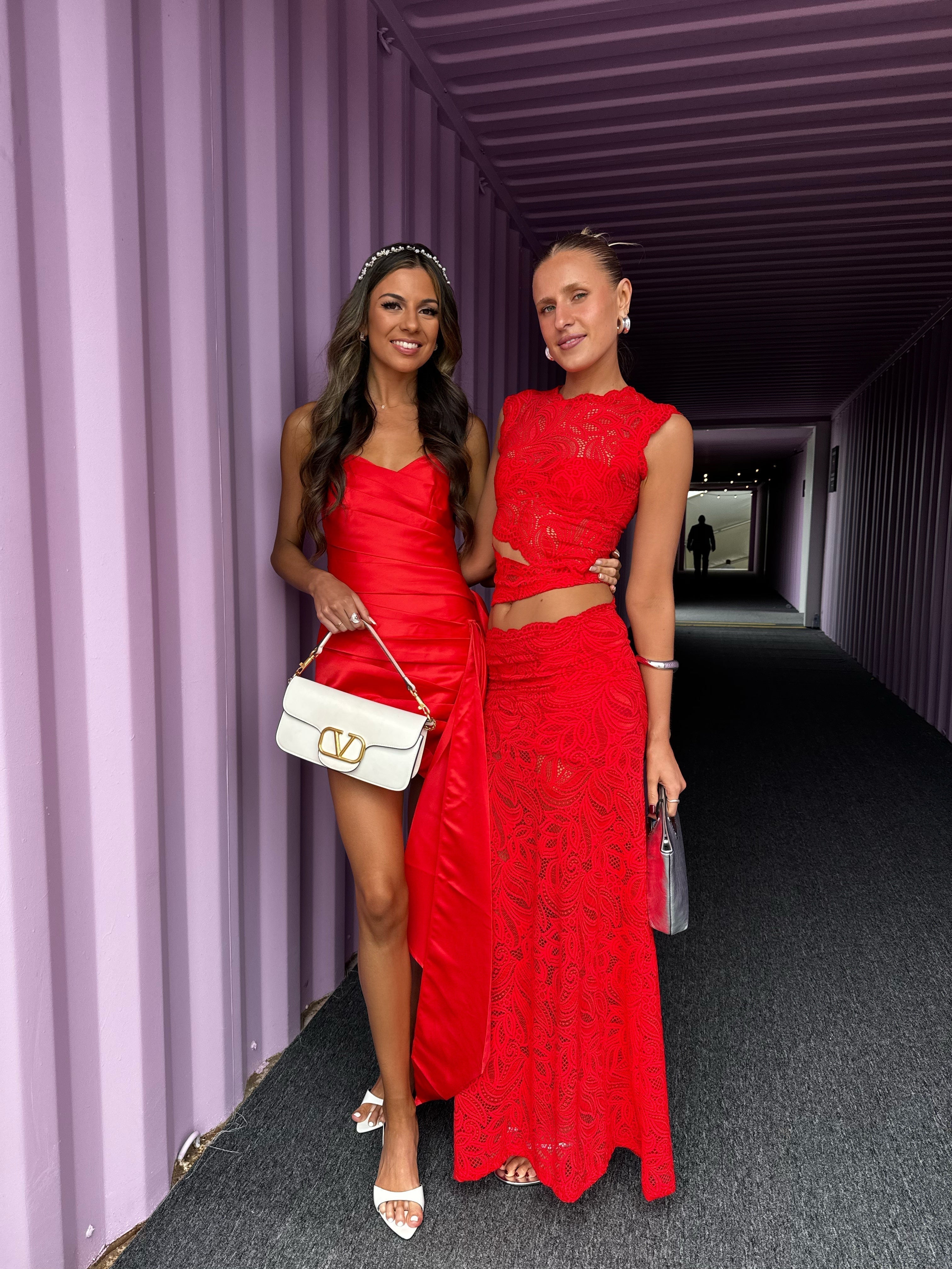 Two women posing, one in a short red dress and the other in a red lace maxi dress set with cutouts.