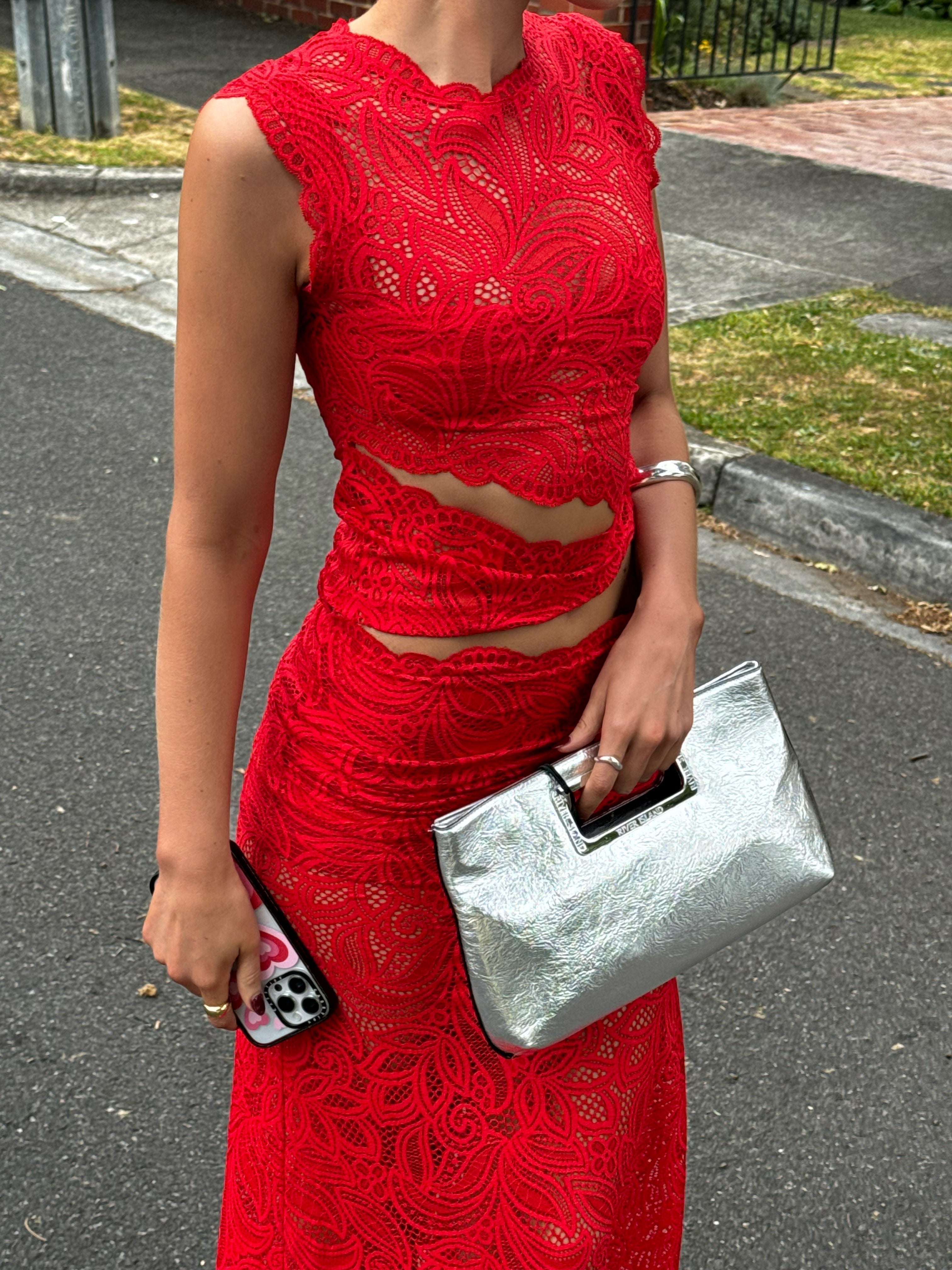 Mid-shot of a woman in a red lace maxi dress set, holding a metallic silver clutch and a phone.