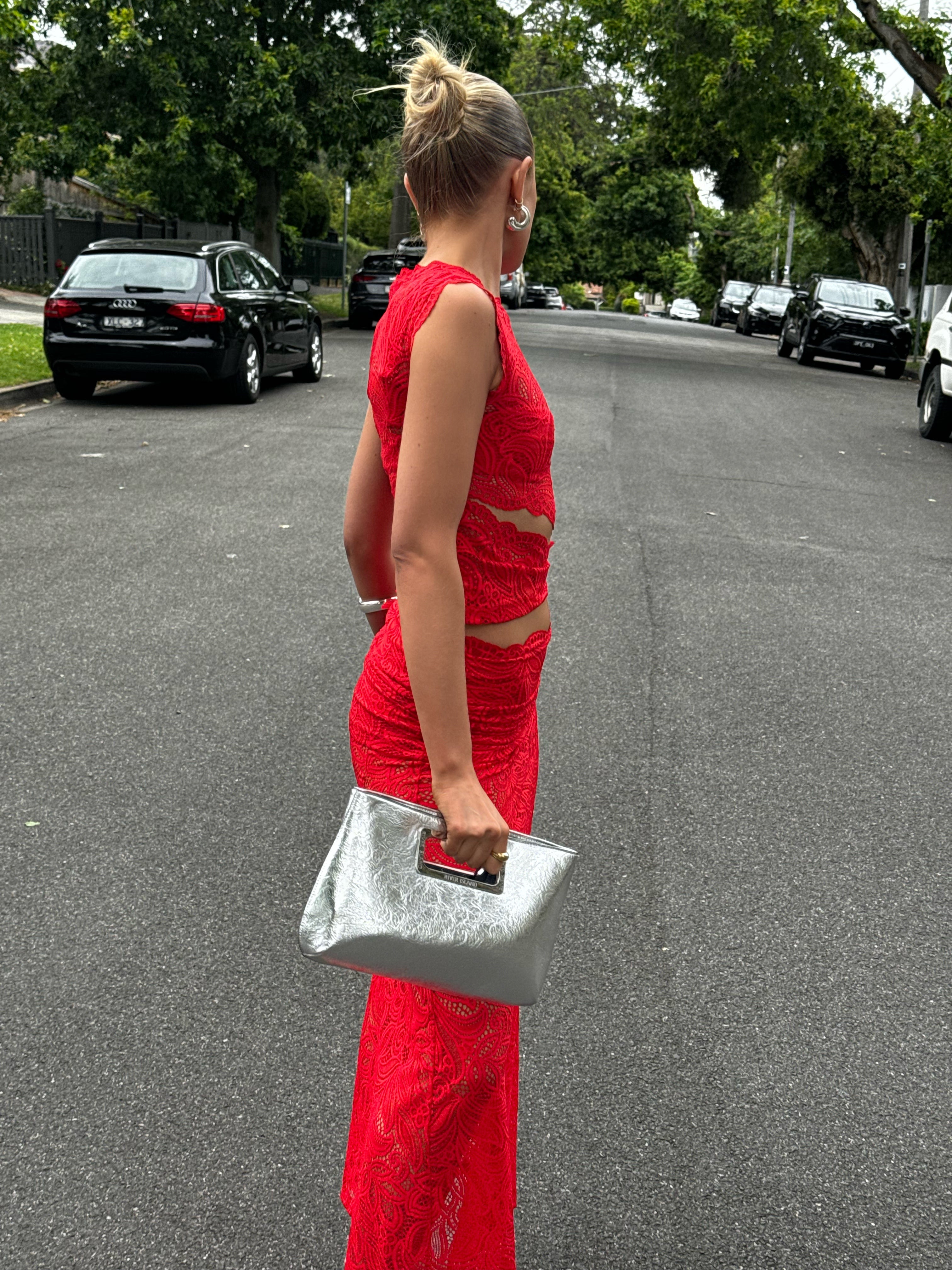 Side view of a woman in a red lace maxi dress set, walking outdoors and holding a silver clutch.