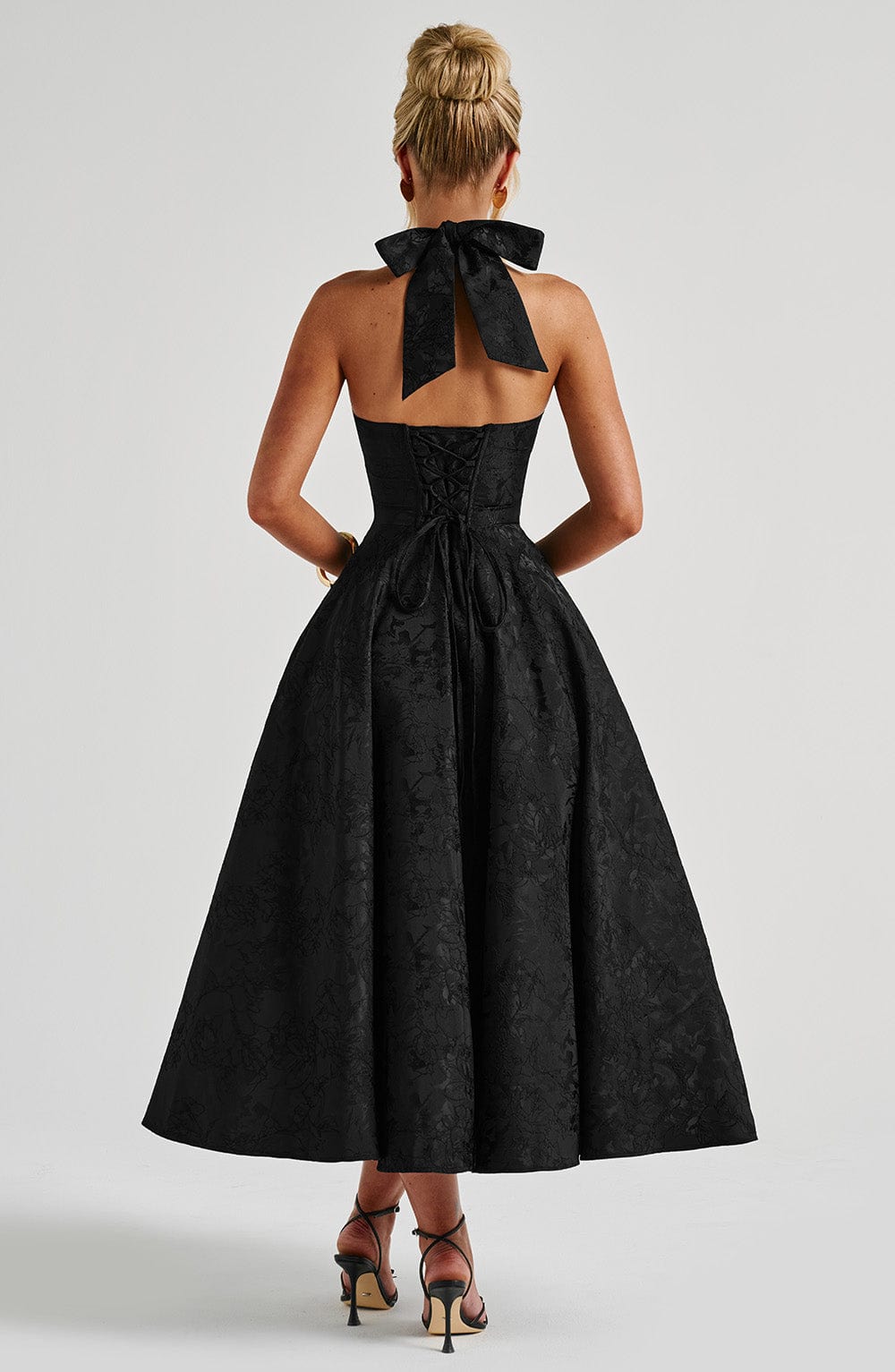 Back view of the black Leilani Halter Midi Dress, showcasing the large halter bow and lace-up corset detail.
