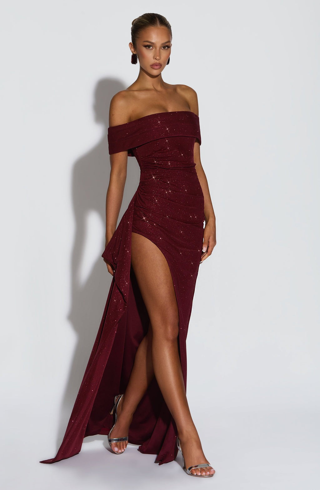 Angled view of a shimmering maroon off-shoulder maxi dress featuring a thigh-high slit and elegant draping.