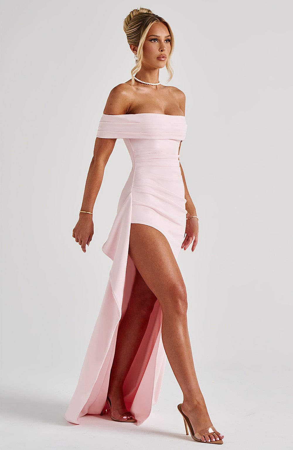 Three-quarter view of a light pink off-the-shoulder maxi dress with a high slit and clear heels