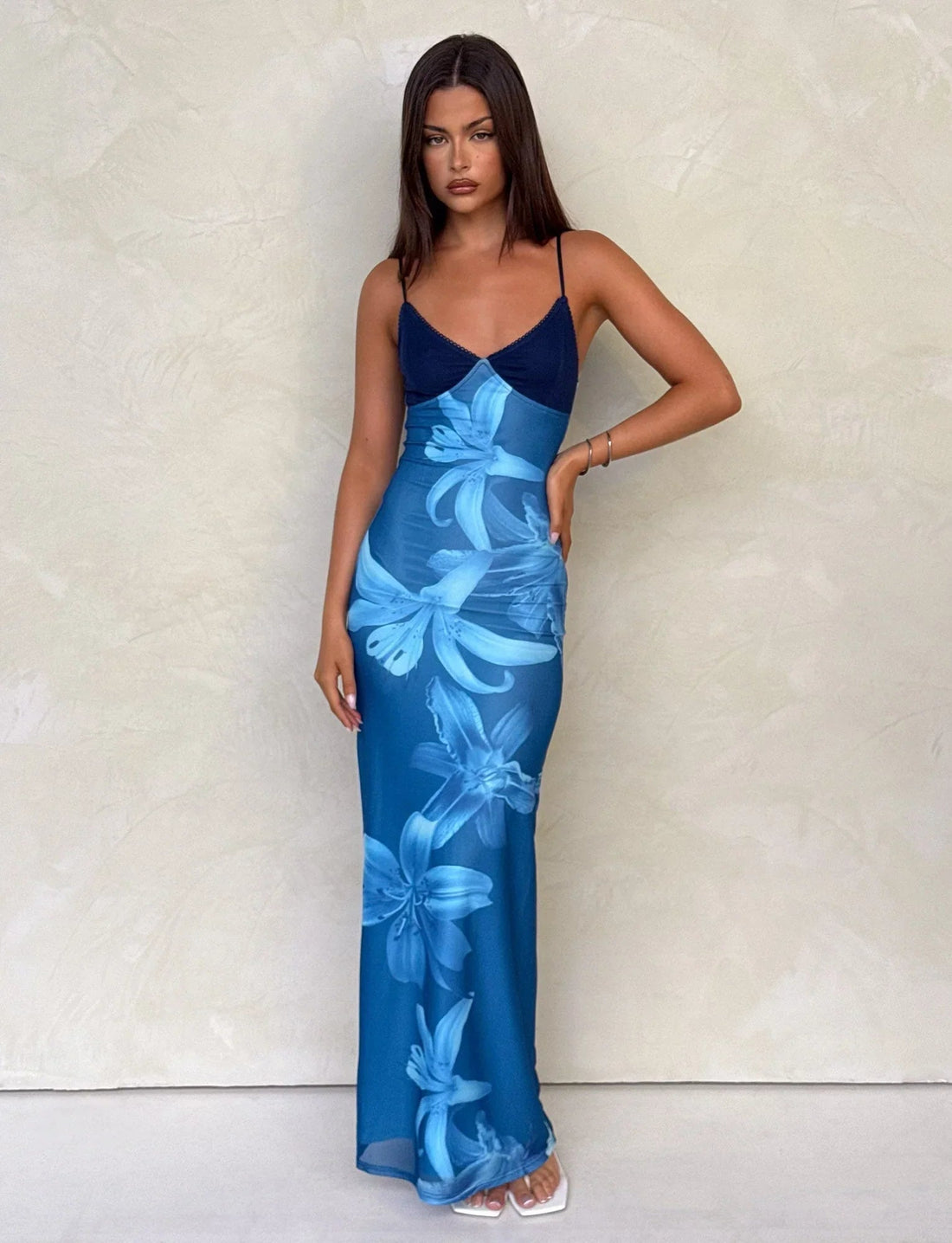 Woman wearing a blue floral maxi dress with spaghetti straps against a light textured wall.